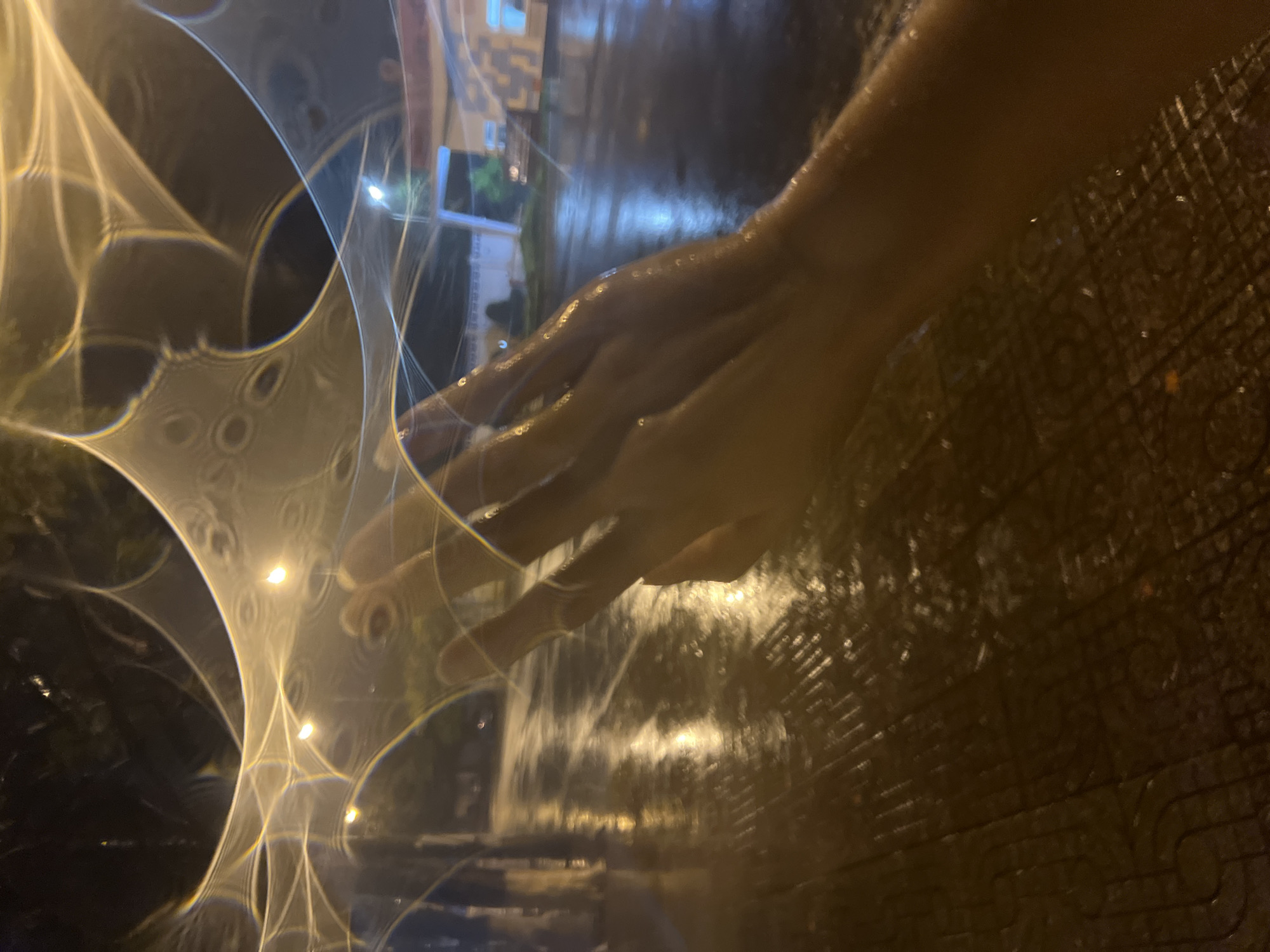 A human hand is positioned over a reflective surface, possibly wet pavement, in an urban setting. There are lights and indistinct shapes visible through a distorted lens or reflective medium. The location is Bac Nha Trang Ward, Vietnam.