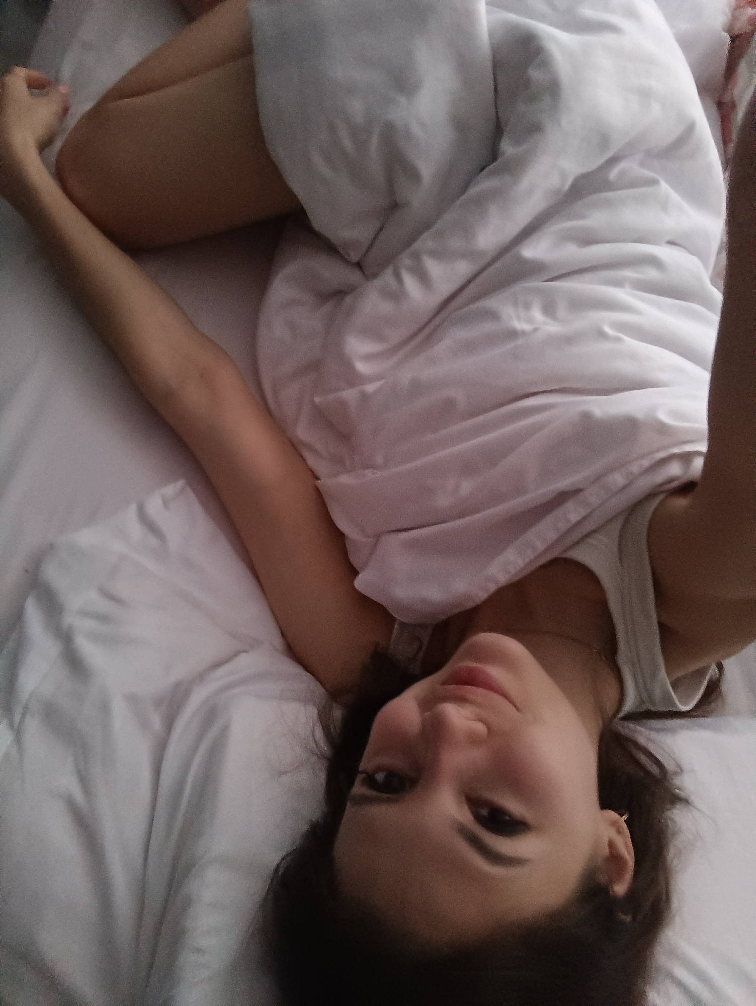 A woman lies in a bed covered with white sheets and a white blanket, her head towards the bottom of the frame. She faces the camera with a slight smile, her dark hair framing her face. She is wearing a white tank top. One arm is outstretched above her head, and her knees are bent. The location is a bedroom in an unknown city.