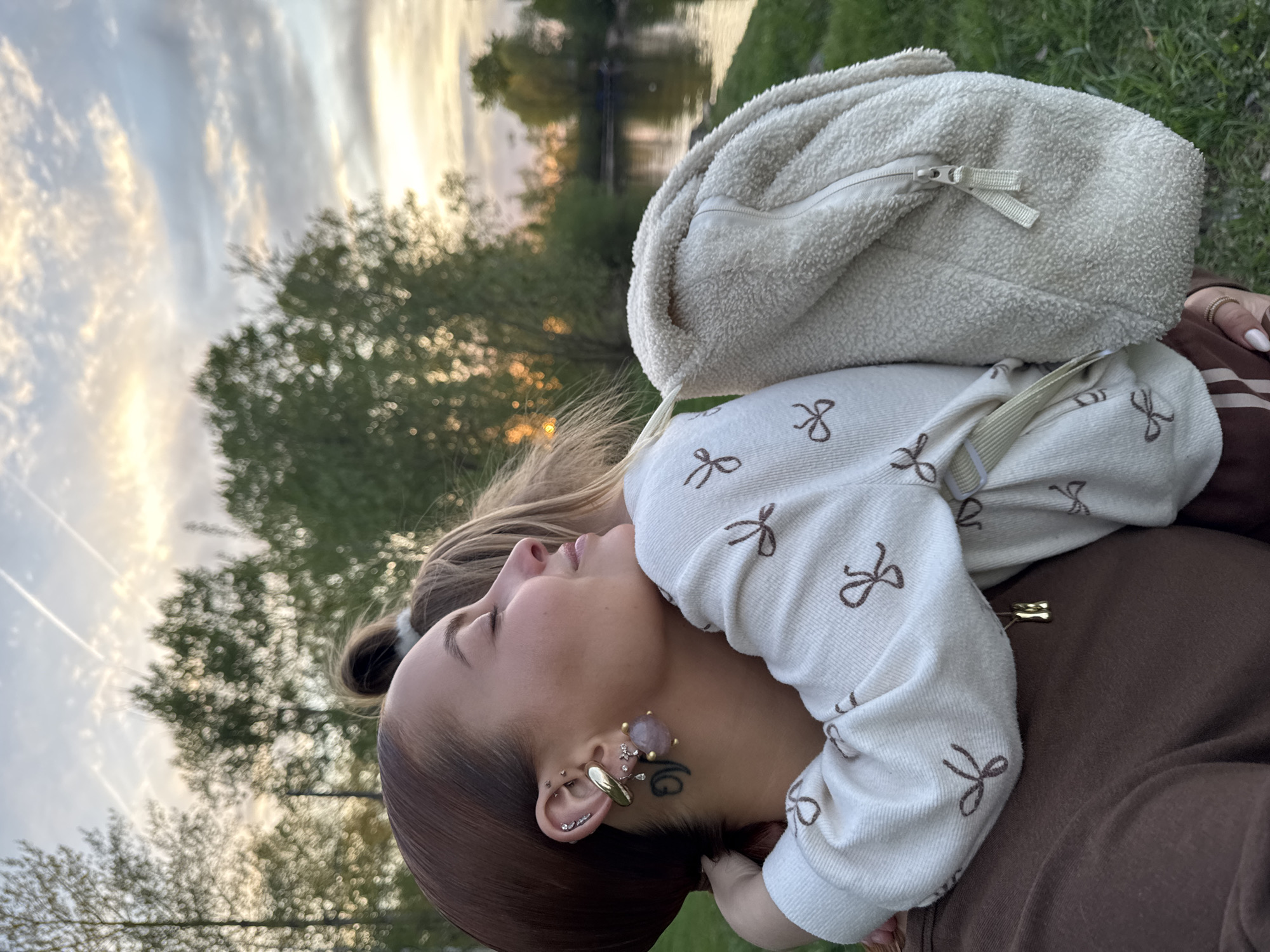 A young woman lies on green grass near a body of water, possibly a lake or wide river, with trees lining the opposite bank. The sky is visible with streaks of clouds reflecting golden sunlight, suggesting sunset. She wears a light-colored sweatshirt with a bow pattern and a brown jumpsuit. A white fluffy backpack rests on her back. She has multiple ear piercings with a variety of earrings, and a tattoo is visible on her neck. She appears to be in a relaxed pose, looking upwards. The location is Chaikovsky, Russia.