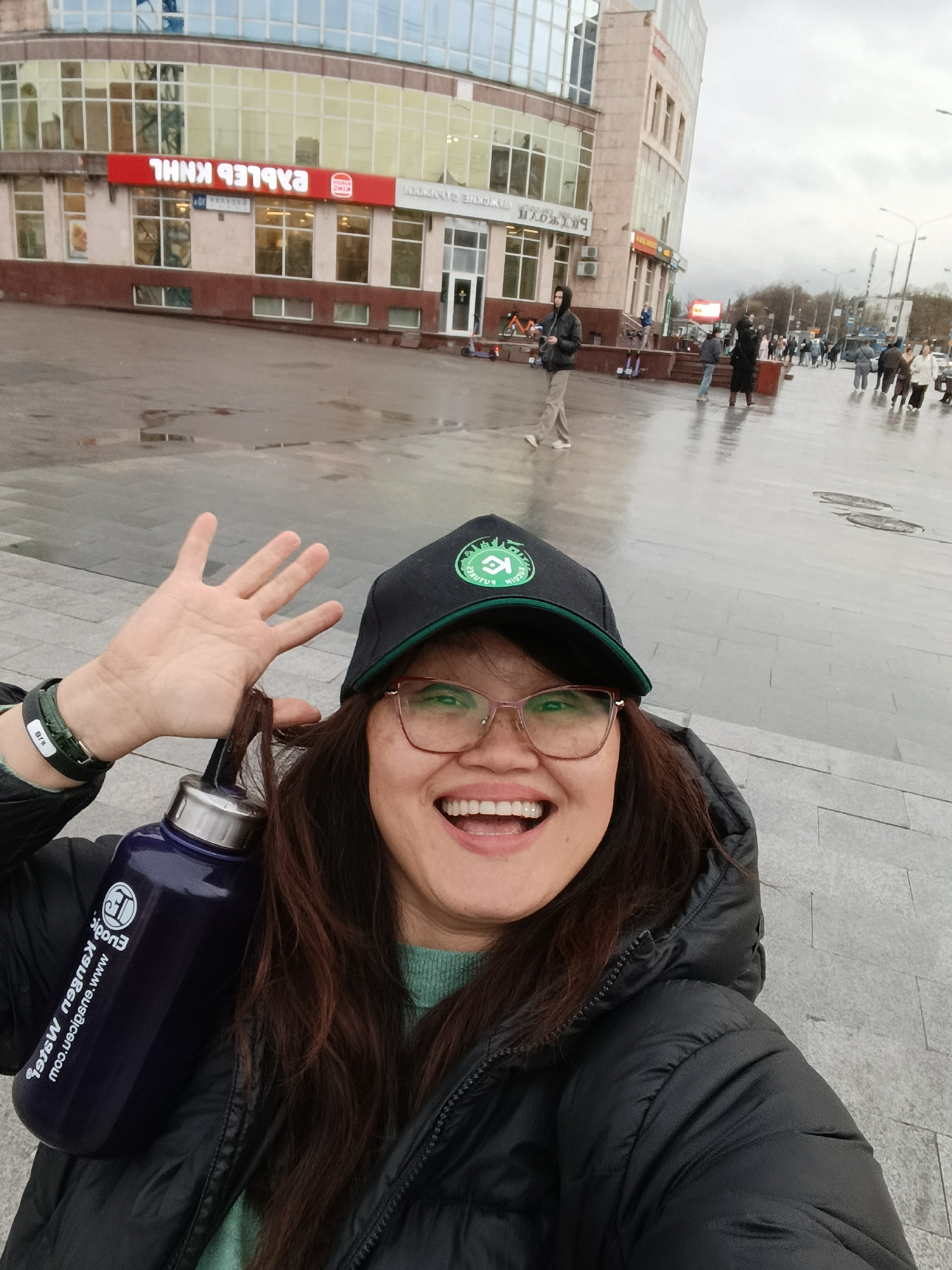 A woman takes a selfie on a cloudy day in Moscow, Russia. She wears a black puffer jacket, green sweater, glasses, and a black baseball cap with a green logo. She is holding up her hand and smiling. In the background, a multi-story building is visible with storefronts; one is a Burger King. The pavement is wet and reflects the sky, and several people are walking in the distance.