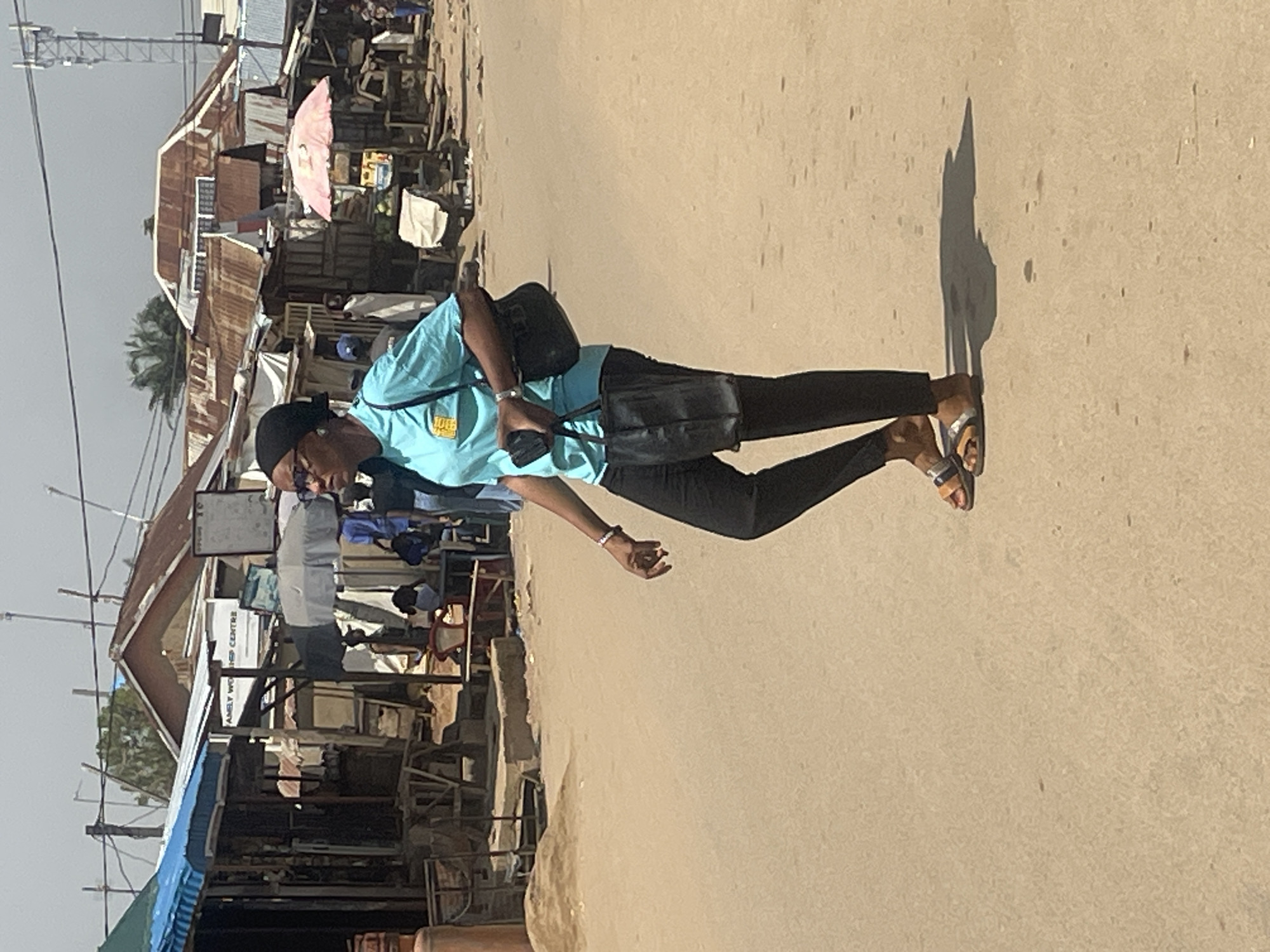 A woman is walking in a dusty, unpaved street in Abuja, Nigeria. She is wearing a light blue t-shirt with a yellow logo, black trousers, and sandals. She has a black headscarf and sunglasses. She is carrying two black bags. One is a purse slung over her left shoulder, and the other is a tote bag in her right hand. The background shows a street market with numerous small stalls made of wood and corrugated metal. Some stalls have awnings and umbrellas. People are visible in the background, some standing and some possibly sitting. Power lines and poles are visible in the sky. The overall impression is of a bustling, informal marketplace.