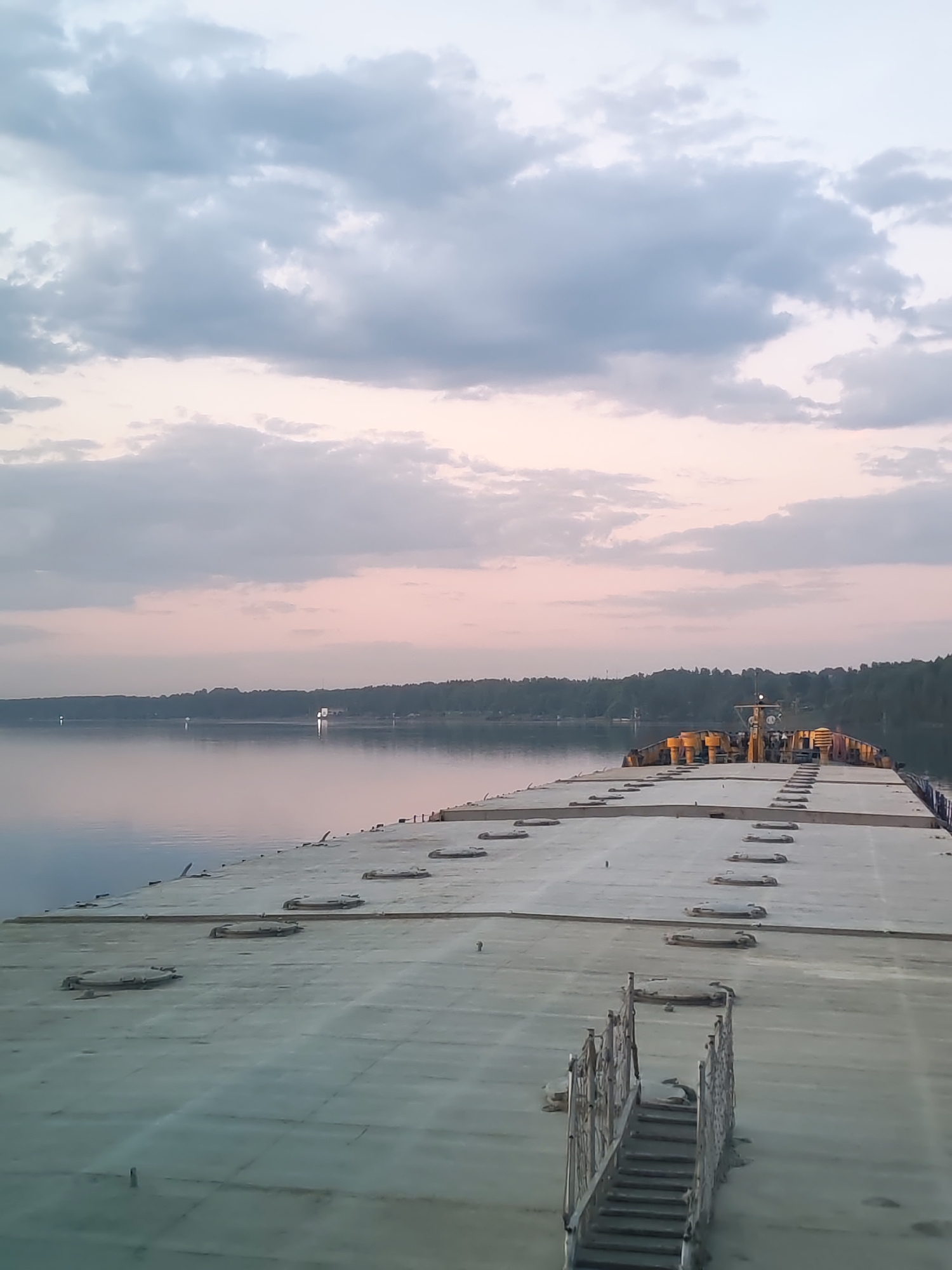 The image depicts the deck of a barge or ship on a body of water, possibly the Protva River near Ermolino, Russia, given the provided location. The deck is flat, light gray, and has several circular hatches or access points. A metal staircase leads down to the deck from the foreground. The water is calm and reflects the light of the sky. A line of trees is visible on the distant shore. The sky is cloudy with hues of gray, white, and pink, suggesting either sunrise or sunset. The lighting is soft and diffused.