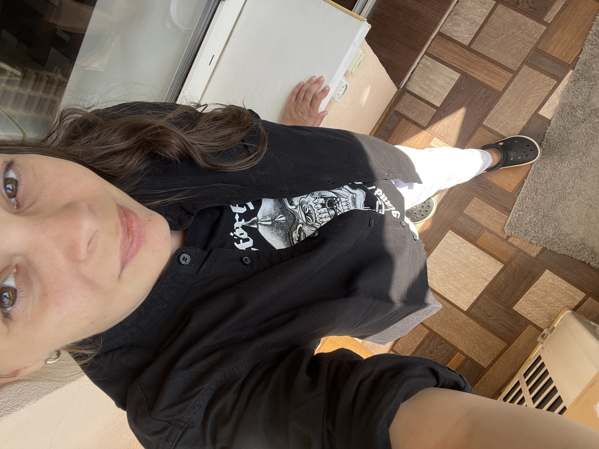 The image shows a first-person perspective of a woman standing indoors, possibly on a balcony or near a doorway, as indicated by the flooring and the presence of a window or glass door. She is wearing a black button-up shirt over a t-shirt with a graphic print, white pants, and black Crocs. The flooring appears to be made of wood-patterned tiles arranged in a herringbone pattern. A light-colored rug is visible on the right side of the image. The background includes a white surface, possibly a cabinet or appliance, and reflections in a glass surface. The scene is lit by natural light, casting shadows on the floor. There is no identifiable information in the image that would suggest a specific location within the city of Tolyatti, Russia.