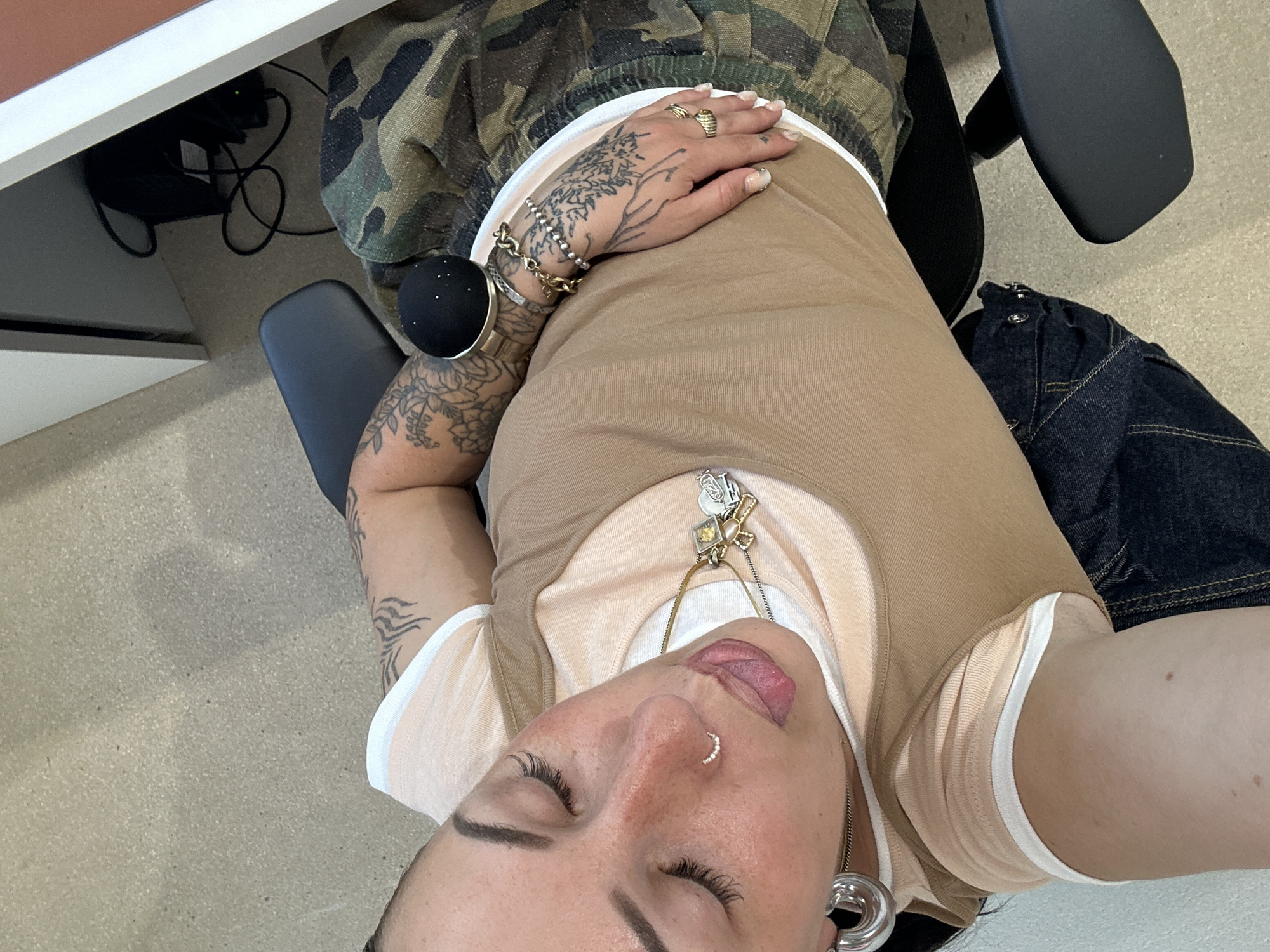 The image is a selfie of a person seated on an office chair with a partial view of a desk on the left. The person has a nose ring, closed eyes, and their tongue slightly sticking out. They are wearing a tan tank top over a lighter peach-colored shirt with white trim. Multiple necklaces adorn their neck. The person has several tattoos on their arms and hand. They are wearing rings and bracelets on their right hand. The person is sitting on an office chair, wearing camouflage pants. A dark denim garment is visible to the right, draped over the chair. There is a black, spherical object on the arm of the chair, near their forearm. A portion of a desk with black cables and equipment is visible in the upper-left corner. The environment appears to be an office setting. The image does not provide any evidence indicating it was taken in Paris, France.