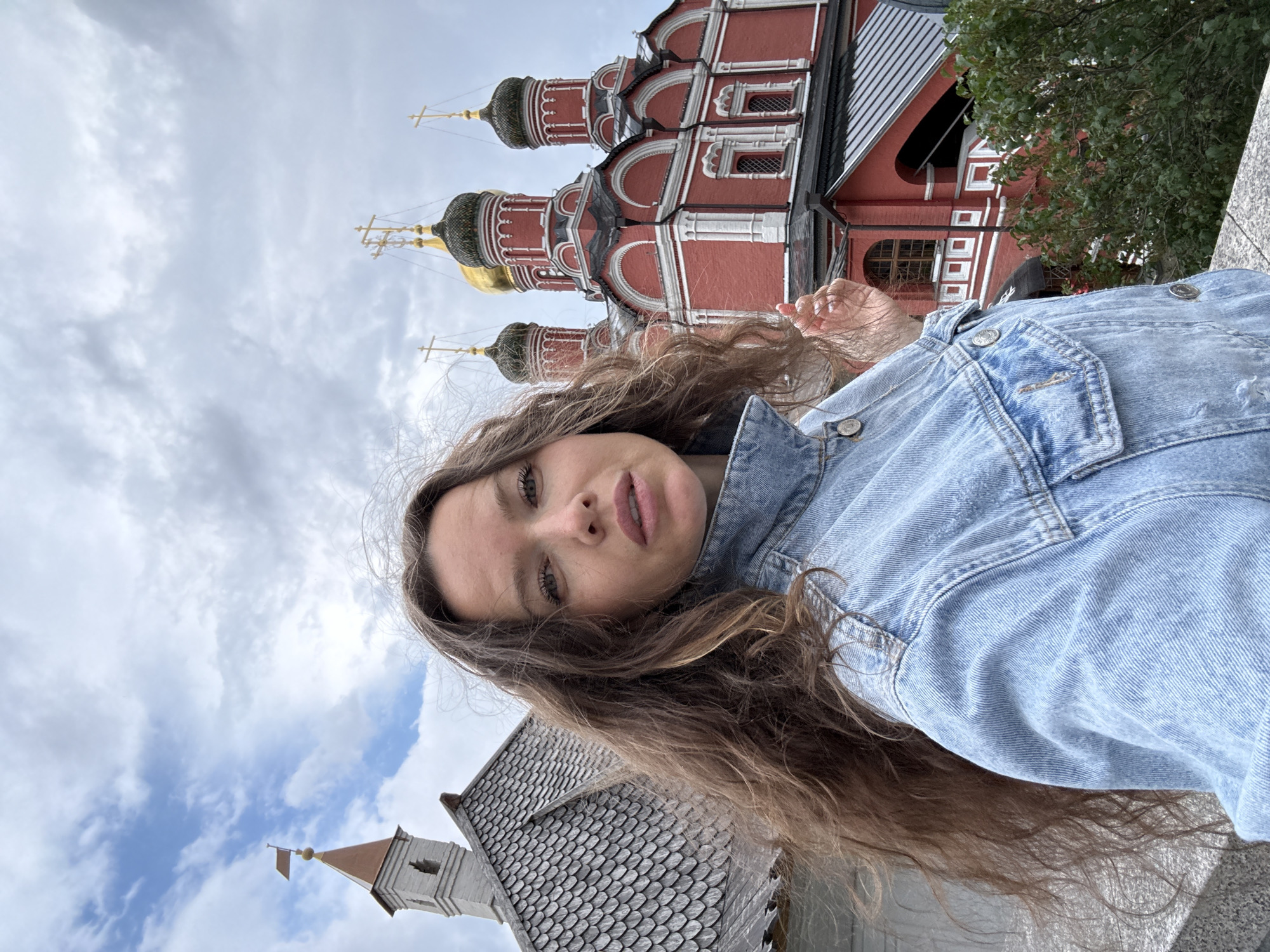 A woman with long, wavy brown hair is taking a selfie with a religious complex in the background. She is wearing a light blue denim jacket. The complex features several red buildings with white trim, rounded windows, and onion domes topped with crosses. One dome is gold. To the left of the complex, a separate, lighter-colored tower with a copper-colored pointed roof and a small flag is visible. The sky is overcast with scattered clouds. The scene appears to be outdoors, possibly on a slightly elevated viewing platform. This location is likely the Cathedral of the Dormition in the Moscow Kremlin, or a similar historical Russian Orthodox religious site.