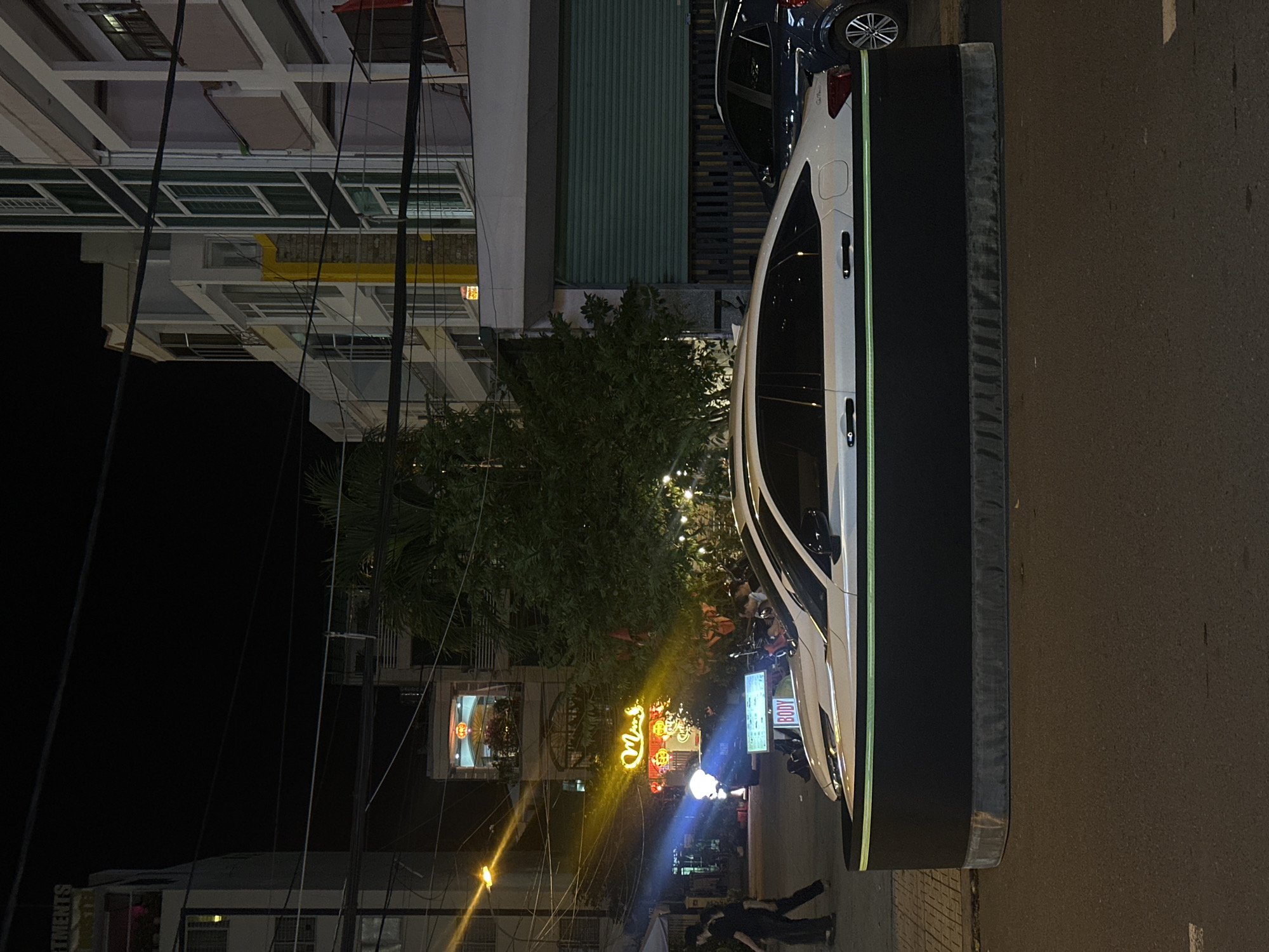 This media file captures a vibrant street scene at night in an urban environment, likely in Nha Trang Ward, Vietnam. The darkness of the sky is illuminated by numerous artificial light sources, including streetlights, building lights, and neon signs, creating a lively atmosphere.
In the foreground, a white sedan is prominently parked on a wide, dark platform or curb that runs parallel to the asphalt road. A thin, glowing green light strip is visible along the side of this platform, adjacent to the car. A darker blue car is parked behind the white one.
To the left, multi-story buildings line the street, showcasing various windows, balconies, and architectural details. Overhead utility wires crisscross the frame, a common sight in urban settings. A large, leafy tree adorned with decorative lights stands in the mid-ground, adding to the nighttime illumination.
Several illuminated signs are visible. A prominent neon sign in the mid-ground displays the Vietnamese word "Mây." Nearby, a rectangular sign clearly shows the text "BODY." In the far background on the left, the partial word "RTMENTS" is discernible, suggesting an "APARTMENTS" sign.
On the dark asphalt road, faint markings can be seen. Towards the bottom right of the image, several people are visible, suggesting ongoing pedestrian activity on the street or sidewalk. The scene depicts a typical, active night in a city, with businesses open and people moving about.