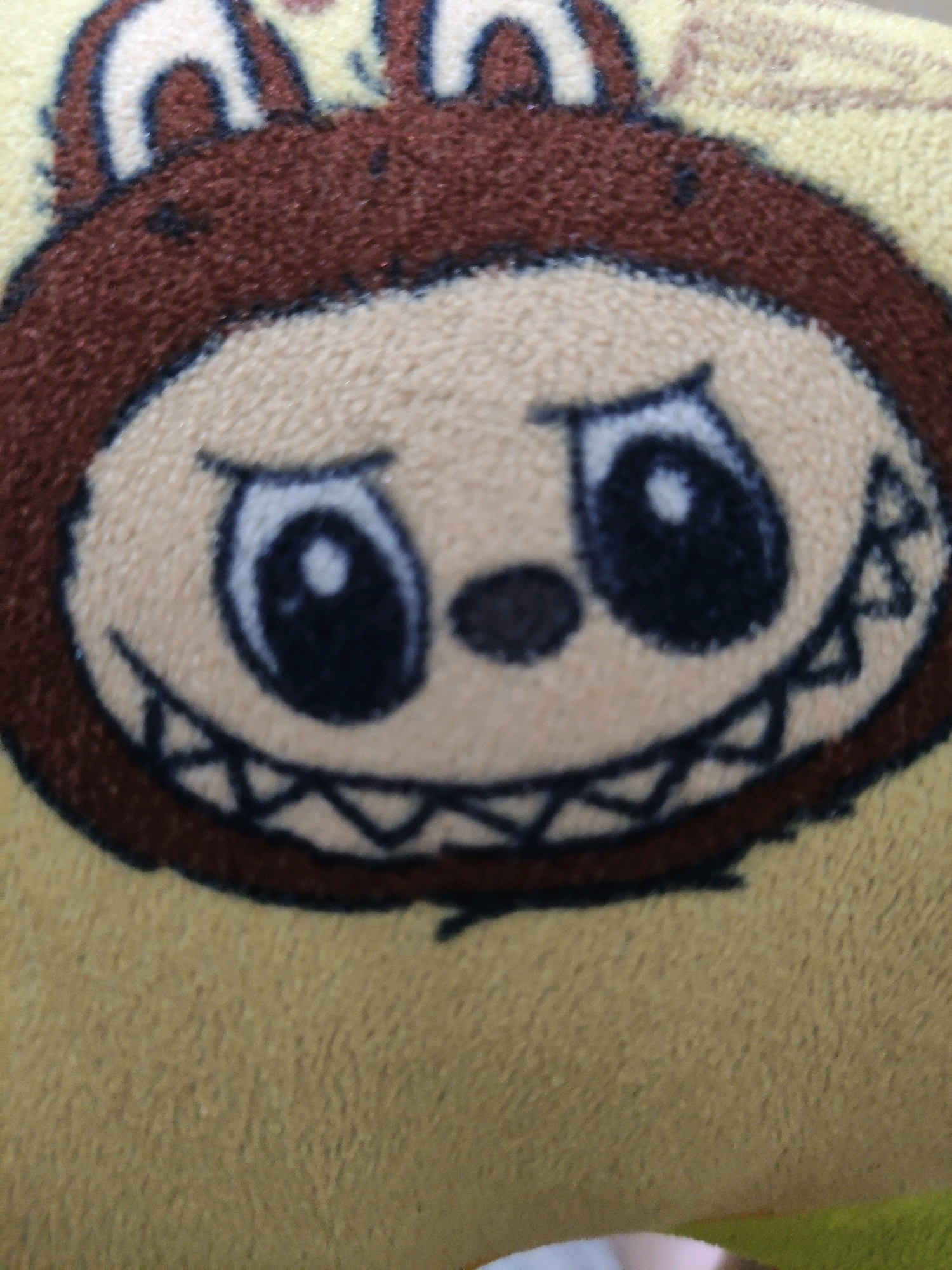 The image shows a close-up of a stuffed toy or plush fabric with a cartoonish bear-like face. The face is round with large black eyes, a small dark nose, and a wide, toothy grin. The toy has brown fuzzy ears and a brown outline around the face. The background is a textured yellow fabric. There is no indication of the location in Bulukumba, Indonesia.