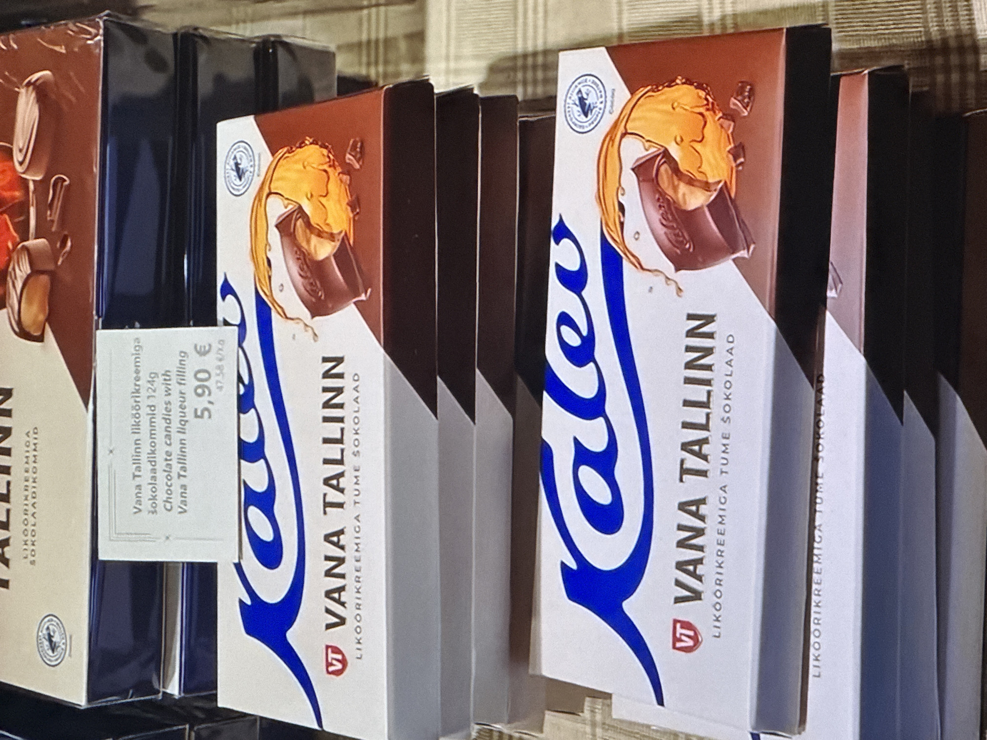 The image shows a stack of Kalev brand chocolate boxes. The boxes are white with blue and brown accents and feature images of chocolate candies with caramel filling. The text on the boxes is in Estonian and English. One box has a price tag that reads "5,90 €". The chocolates appear to be in a retail setting, possibly a store in Tallinn, Estonia, given the brand name "Vana Tallinn" which is a well-known Estonian liqueur and chocolate brand. The lighting suggests an indoor environment with artificial light.