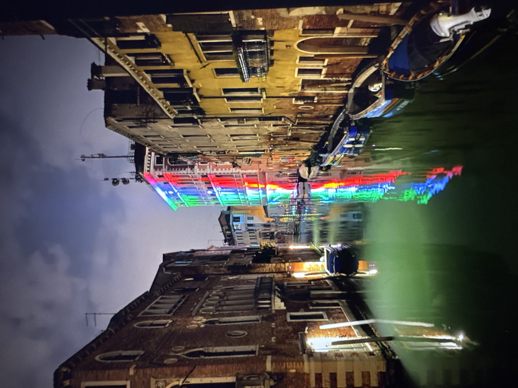 The image depicts a canal scene at night, with buildings lining both sides and boats moored along the waterways. The canal reflects the lights from the buildings and the sky, which is filled with clouds. One building, in the distance, is illuminated with colorful lights that create a vibrant spectrum on the water. Several boats are visible, some empty and others with what appear to be people inside or objects covered. The architecture suggests an old European city. The water is a dark green color. The scene is dark and atmospheric due to the nighttime setting. There is no indication that this location is in Porrentruy, Switzerland.