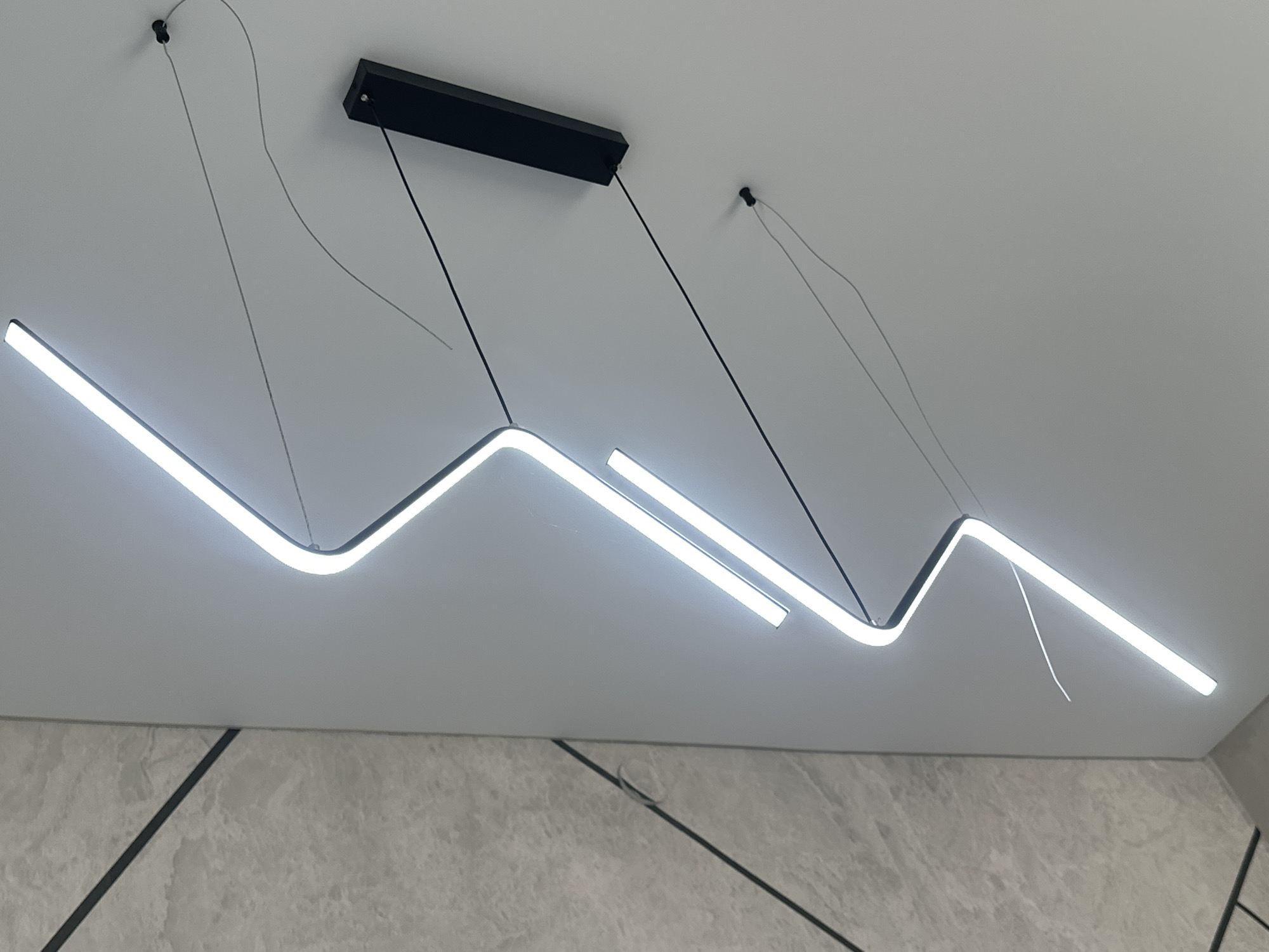 The image shows a modern interior with a white wall and a grey, textured wall on the left. A zigzagging LED light fixture is mounted on the white wall. A black rectangular electrical junction box is also visible on the white wall. The scene is illuminated by the LED light. The location in the city of Tabuny, Russia is not discernible from the image.