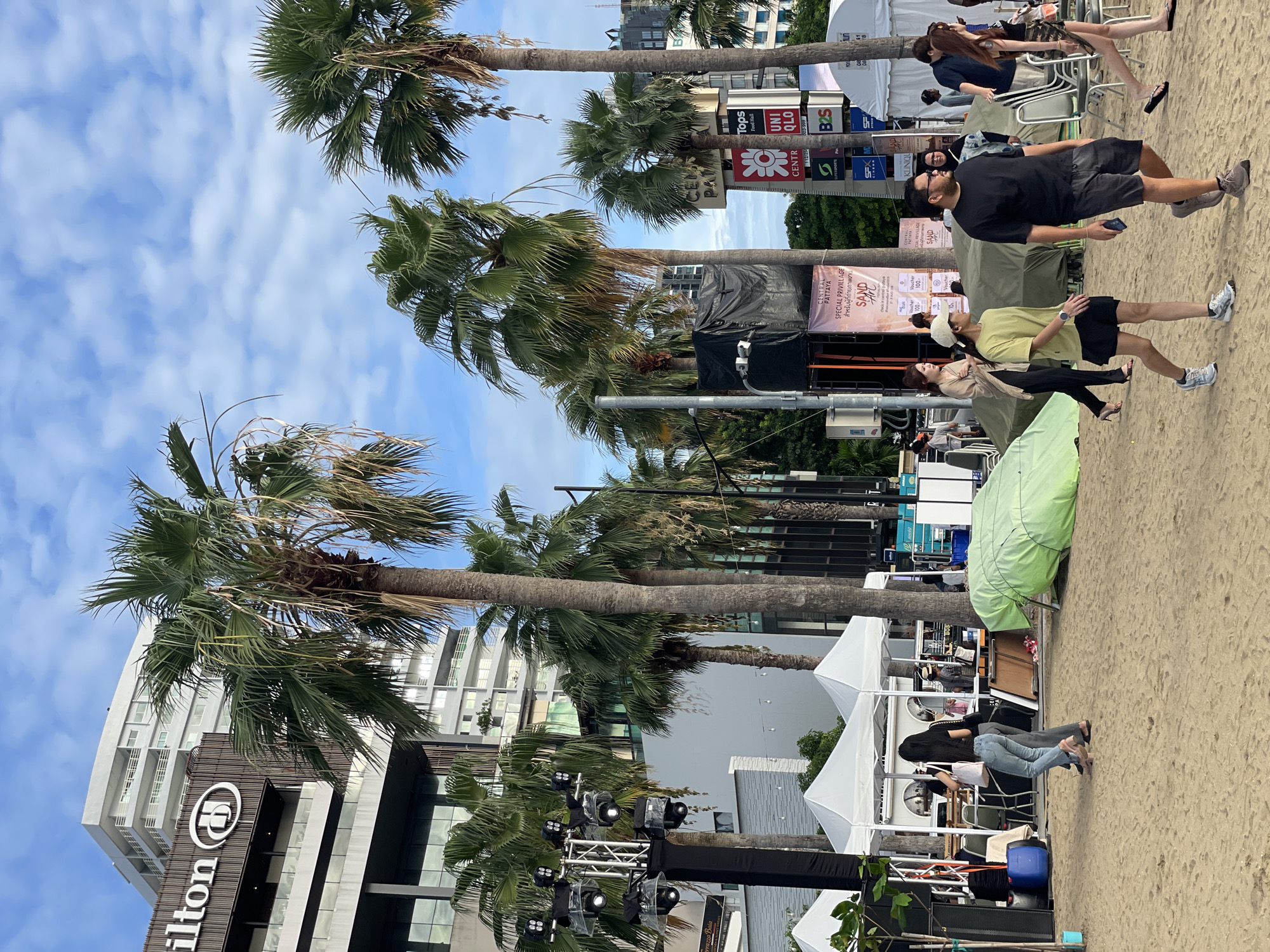 The image captures a scene on a sandy beach in Pattaya City, Thailand, with the Hilton Pattaya hotel prominently visible in the background. The beach area is set up with temporary structures, including white tents, likely for vendors or an event. Palm trees are scattered throughout the scene, typical of a tropical coastal environment. Several people are walking on the sand, dressed casually in warm-weather clothing. One group is near the tents, with some individuals appearing to browse or interact with the stalls. Another group is walking further down the beach, further away from the camera. In the mid-ground, there is a stage setup with lighting equipment and black tarps, suggesting a performance or event is either about to happen or has recently concluded. The sky is partly cloudy, indicating an overcast but potentially bright day. Signs for businesses like "Tops Food Hall," "UNIQLO," and "CENTRAL PATTAYA" are visible on a pole in the background, identifying the area as a commercial district adjacent to the beach. A green tarp is covering an object on the sand, possibly equipment or seating.