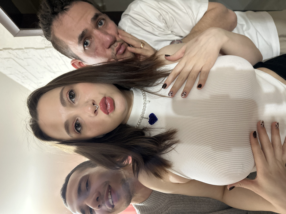 The scene depicts three individuals, one female and two males, posing closely together in what appears to be an indoor setting in the city of Saratov, Russia. The central figure is a female with long, dark brown hair, wearing a white ribbed sleeveless top, a pearl choker necklace with a dark blue heart-shaped pendant, and a silver septum ring. She has visible makeup, including glossy lips and glitter eyeshadow, and her fingernails are painted with both black and leopard print designs. She is puckering her lips and directly facing the camera, with both hands placed on her chest just below her breasts. To her left and slightly behind her is a male with short, dark hair, wearing a light grey knitted sweater, who is smiling and looking towards the camera. To her right and also slightly behind her is another male with dark, possibly dreaded hair, wearing a white t-shirt. This male has his right hand resting against his chin/cheek and wears a gold-colored ring on his right ring finger; he is looking towards the camera with a neutral expression. The background consists of a white wall, partially textured with a brick-like pattern on the right, and a dark brown doorframe is visible on the far right. The lighting is bright, with a reddish hue present in the upper left background.