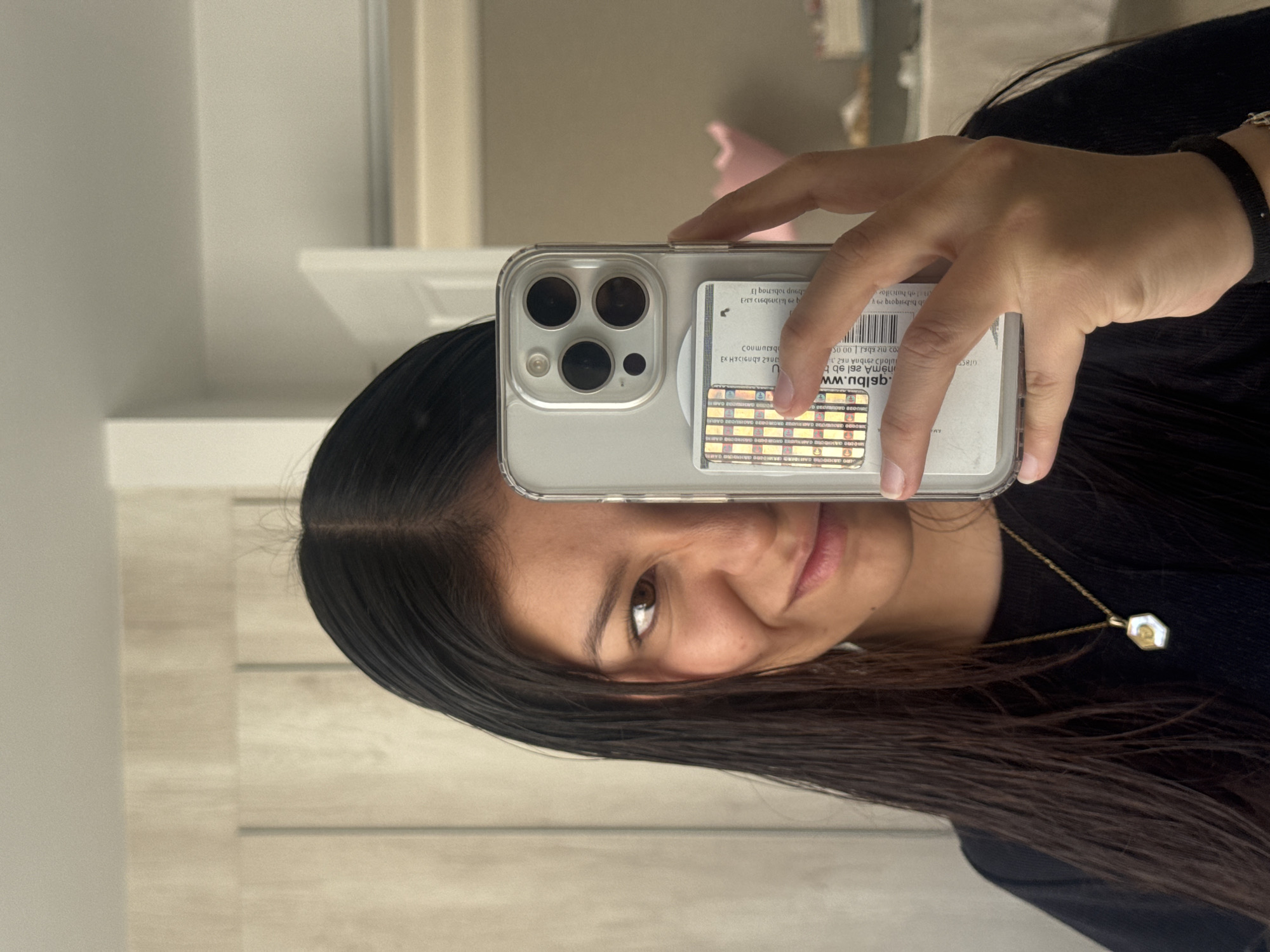 A young woman is captured in a mirror selfie, holding an iPhone with her right hand. She has long, dark hair with a center part, and a subtle smile on her face. She is wearing a dark long-sleeved top and a delicate gold necklace with a light-colored, possibly mother-of-pearl or shell-like, geometric pendant. A dark band or bracelet is visible on her left wrist.
The smartphone appears to be an iPhone 13 or 14 Pro/Pro Max model, identifiable by its clear case and the distinctive triple-lens camera module. A colorful, checkered card holder or grip is attached to the back of the case, and a white card or label is visible beneath the clear case.
The photo is taken indoors, likely reflecting off a mirror or shiny surface, revealing light-colored walls and a light-toned wooden floor in the background. The general lighting suggests it is daytime. This image is set in San Francisco Ocotlán (Ocotlán), Mexico.
The text on the card inside the phone case is reversed due to the mirror reflection. Visible elements include the letter "U", what appears to be a reversed URL fragment like ".www.blpu", the number "50.00", and a barcode. Other mirrored words and phrases such as "El bourgoi," "dneq," "Coumnesq," and "EX HICIGUQY 2900" are also present but difficult to fully decipher in their reversed state.