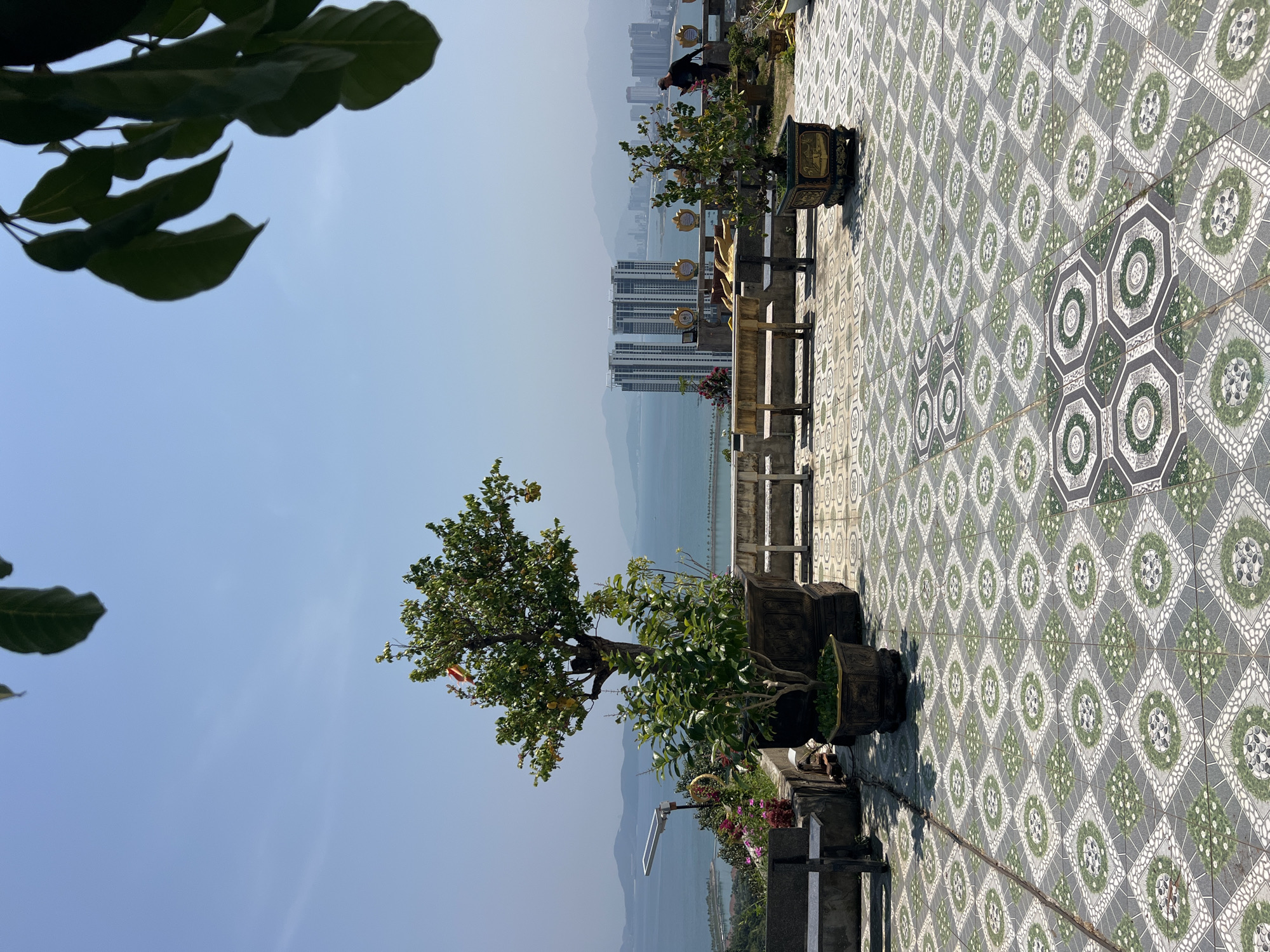 The image shows a view from a high vantage point, likely a temple or elevated platform, overlooking the city of Nha Trang, Vietnam, and the sea. In the foreground, there's a tiled patio with a decorative pattern. Several potted plants and trees are placed along the edge, partially obscuring the view. A figure, possibly a person, is seen in the middle ground, partially hidden by foliage. Tall buildings are visible in the distance, along with mountains fading into the horizon. The sky is clear and blue.