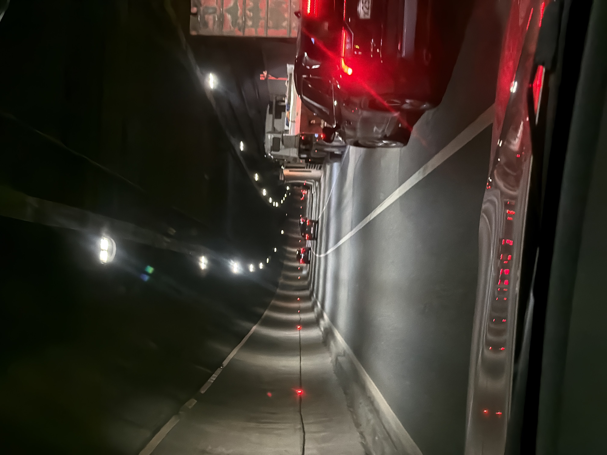 The image shows a traffic jam inside the Lefortovo Tunnel in Moscow, Russia. The tunnel is dimly lit by overhead lights and red marker lights along the road. Cars are visible, with brake lights illuminated, indicating slow or stopped traffic. The walls of the tunnel appear gray and smooth. A partial view of a vehicle is visible to the right.