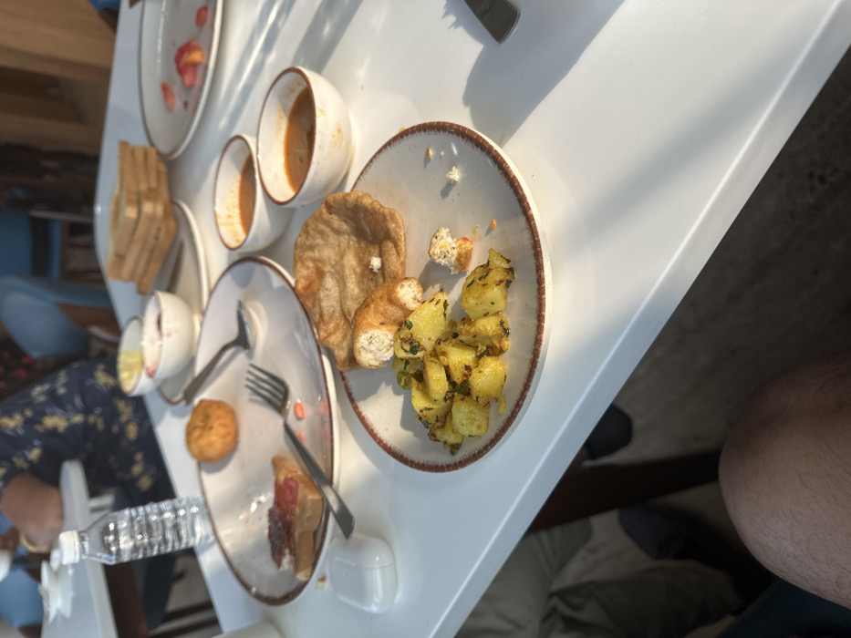 An overhead shot reveals a partially consumed breakfast spread on a white table. Plates contain remnants of various items like bread, puri, potato curry, and pastry. Cutlery, including a fork and spoon, lies on a plate with a small muffin. White ceramic cups and bowls, some stained with remnants of food, add to the scene. A water bottle is visible, while a leg is visible on the side.