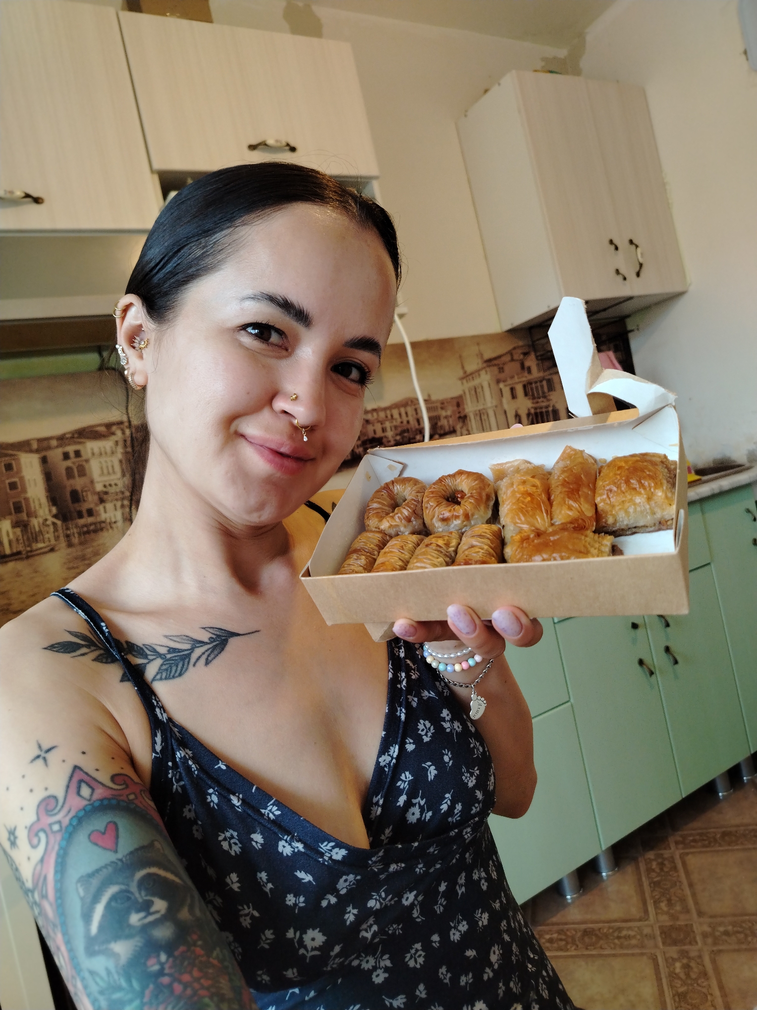 A woman is holding a box of pastries in a kitchen. The woman has dark hair pulled back, multiple piercings in her left ear, and a nose ring. She has a tattoo of a branch on her left shoulder and a colorful tattoo on her left arm. She is wearing a dark blue floral print dress. The box contains several pieces of baklava and other round pastries. The kitchen has white and light green cabinets. The wall behind the pastries has a mural of an Italian cityscape. The floor is tiled in a brown pattern. The city of Noviy, Russia is not mentioned or visibly relevant in the image.