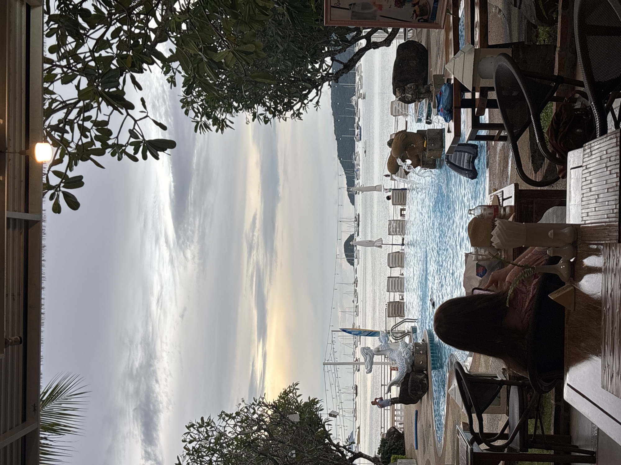 The image depicts a poolside restaurant overlooking a bay filled with sailboats. In the foreground, a person sits at a table with their back to the camera, looking at a phone. The restaurant has outdoor seating with woven chairs and wooden tables. A swimming pool with clear blue water is visible, featuring decorative statues of a mermaid and other figures. The background shows a cloudy sky with the sun setting or rising, casting a warm glow on the water. The location is identified as Chalong, Thailand.