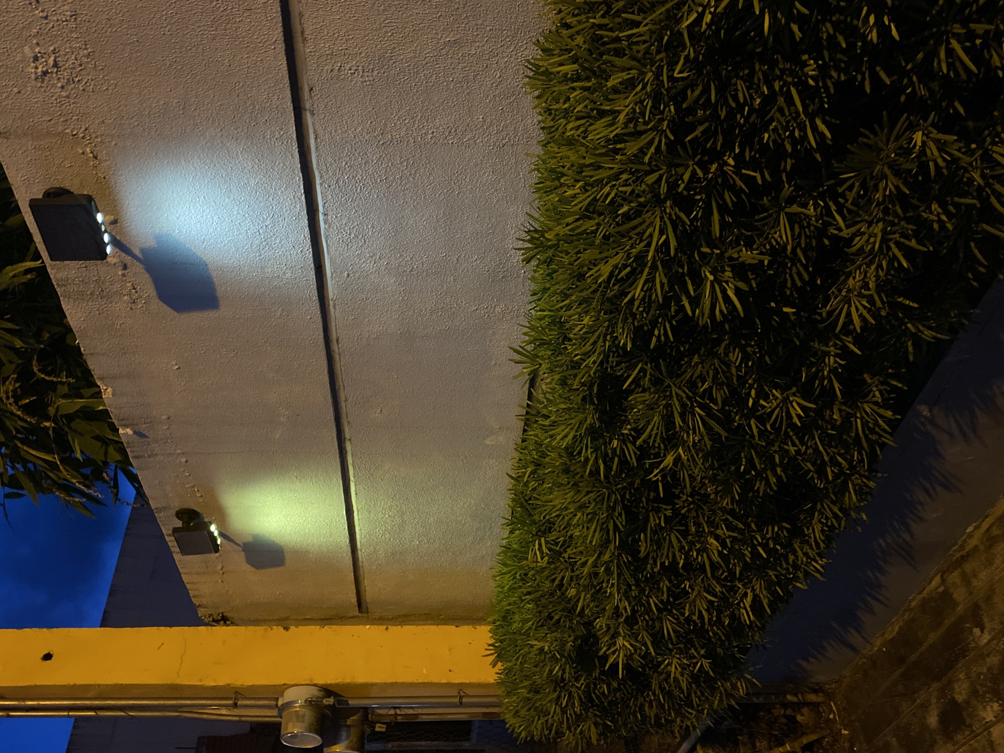 This is a nighttime photograph taken at the perimeter wall of a property in Davao City, Philippines. The image shows a textured, light-colored concrete wall with two modern outdoor wall-mounted lights. The light on the left emits a yellowish hue, while the one on the right casts a cooler, bluish-white light. Both lights illuminate sections of the wall and cast distinct shadows. Below the wall, a dense row of dark green foliage, likely a hedge, runs horizontally across the lower part of the frame. To the far left of the image, a portion of a bright yellow painted post is visible, with what appear to be utility meters or conduits attached to it. The background is a dark blue, indicating a clear night sky. The overall scene depicts an exterior residential or commercial property at night, with artificial lighting illuminating the wall and surrounding vegetation.