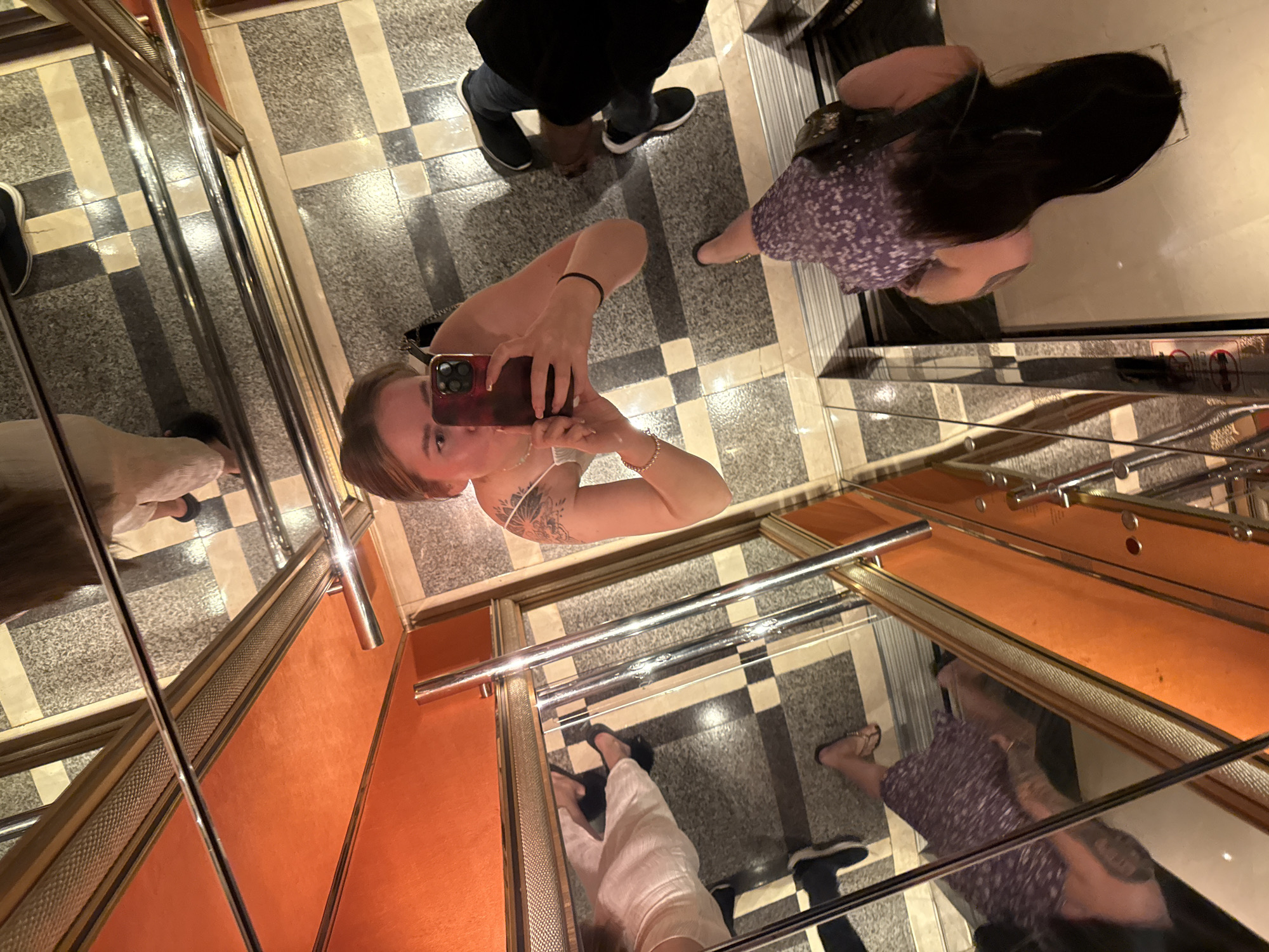 The image depicts the interior of an elevator with a mirrored back wall, creating multiple reflections of the occupants and the elevator's features. The elevator has an orange interior panel. The floor of the elevator has a checkered pattern. A person is taking a selfie in the elevator. Additional people are standing nearby. The exact location of the elevator within the Shangcheng District in China is not discernible from the image.