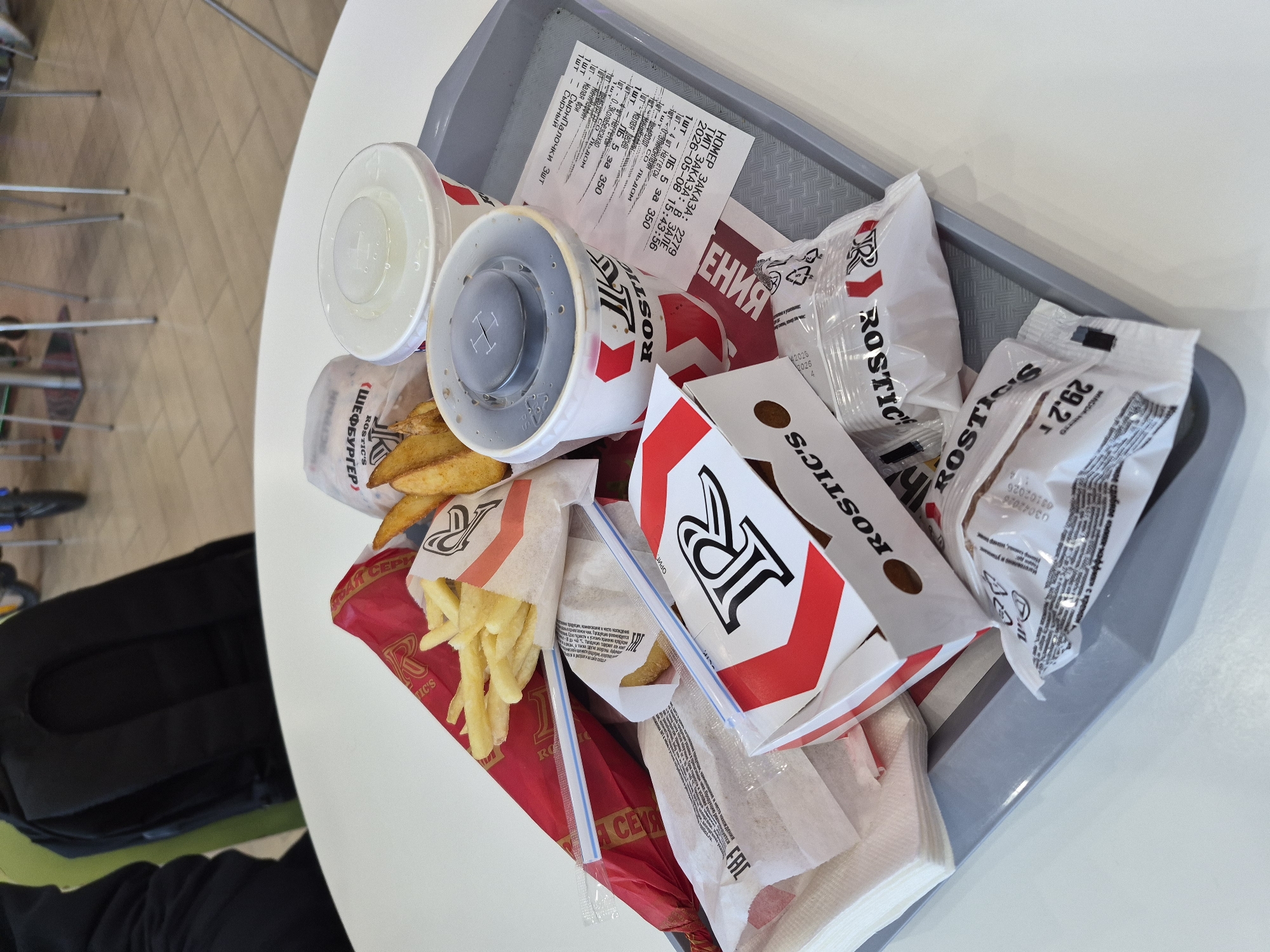 This is a close-up shot of a fast food meal on a gray plastic tray, seemingly from a restaurant chain called "Rostic's". The tray is placed on a white table, likely inside the restaurant. The meal includes:
* Two beverages in red and white cups, one with a grey lid.
* A burger packaged in white paper with branding.
* At least two portions of fries in red and white branded packaging.
* Packets of sauce and napkins.
* A receipt showing the order details.
The receipt reveals the order was placed on May 8, 2026, at 15:43:56 (3:43 PM) inside the restaurant. The meal includes "LB 5" (likely a specific burger or menu item), nuggets, a small serving of fries, a cheeseburger, and cola without sugar. The text on the packaging indicates that the restaurant chain "Rostic's" operates in Tobolsk, Russia. A small portion of a person wearing black clothing is visible at the lower left of the image. Part of a bag, possibly a backpack, is visible, too. Portions of furniture like chairs are visible. The background is neutral.