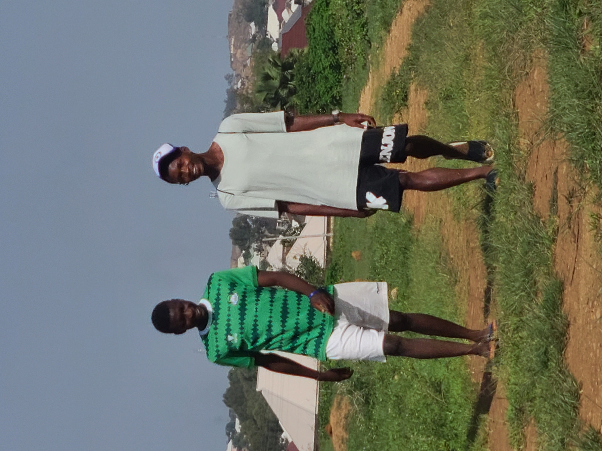 Two men stand outdoors in Unknown, Unknown. The man on the right wears a light gray short-sleeved shirt, black shorts with white lettering, a white baseball cap, and dark sandals. The man on the left wears a green and white short-sleeved shirt with white shorts and colored sandals. They are standing on a grassy area with a dirt path. There are houses and vegetation in the background. The sky is overcast.