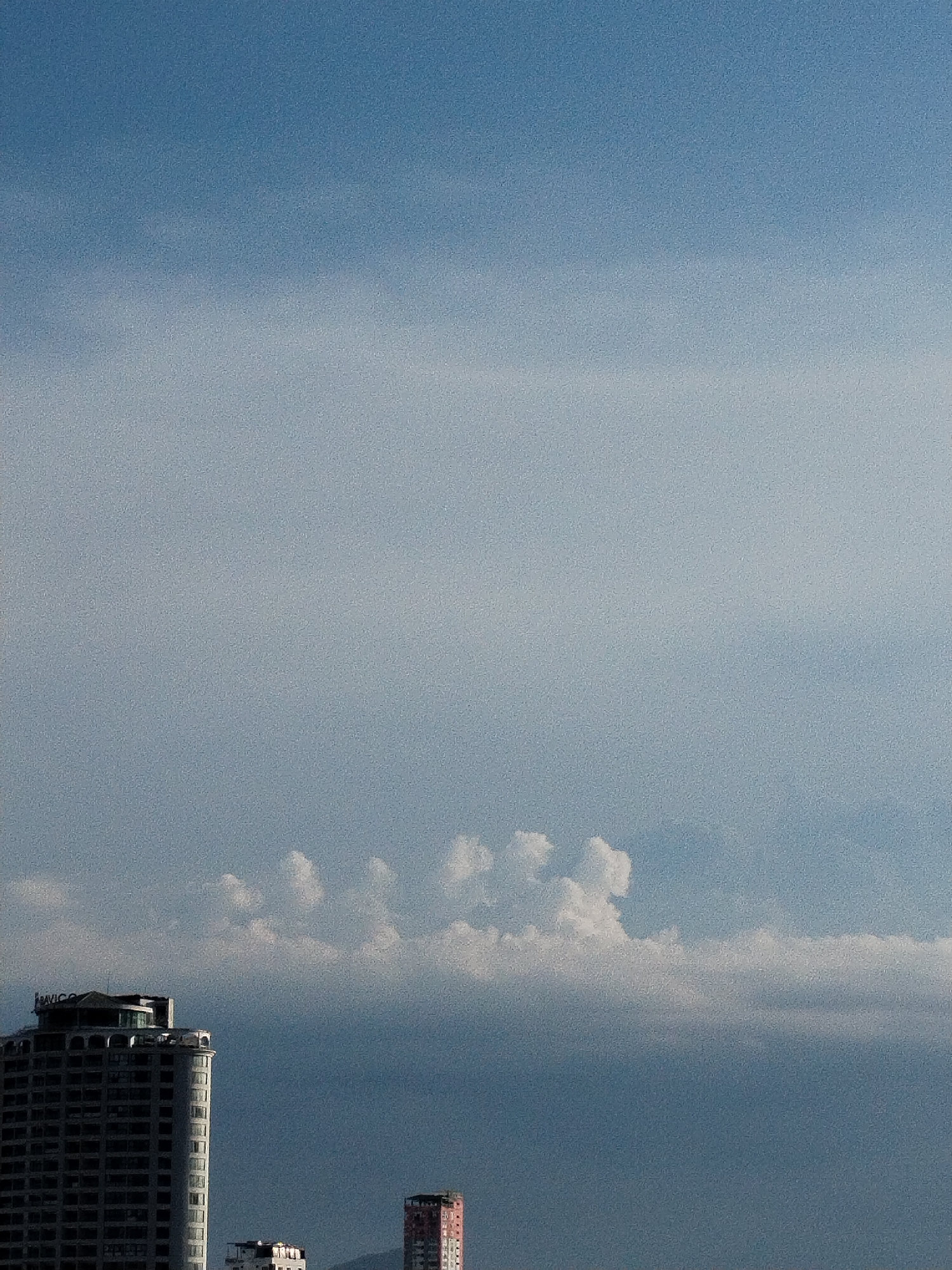 The image shows a cityscape in Nha Trang Ward, Vietnam. In the foreground are three buildings: a tall, gray, multi-story building labeled "AVICO" on its roof, a smaller, white building, and a taller, pinkish-red building. In the background, a cloud formation resembles a row of tufted or fluffy shapes hovering over a hazy, grayish expanse, possibly a mountain range or a large cloud bank. The sky above is a gradient of blue.