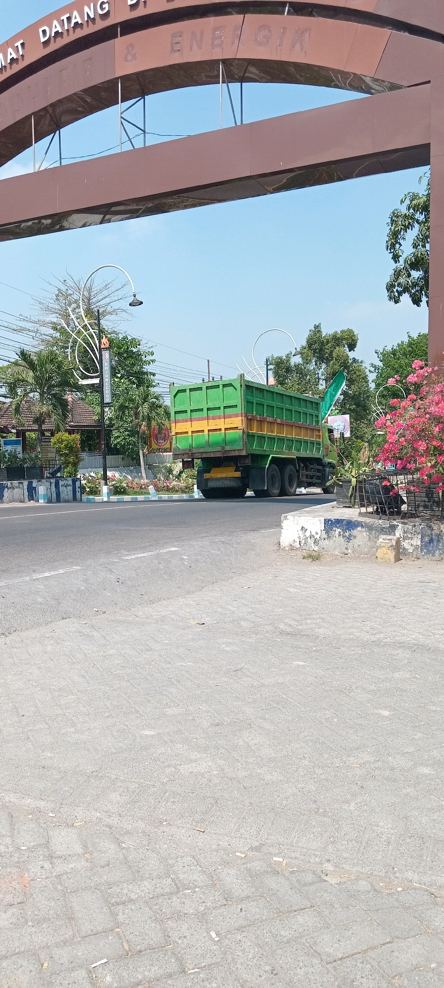 The image shows a street scene in Tulungagung, Indonesia. An arched gateway with white lettering reads "SELAMAT DATANG DI & ENERGI". Beneath the gateway, a green dump truck with a yellow stripe is driving on the right side of the road. The truck has six wheels. To the left of the road, there is a lamppost with decorative white swirls and a sign that says "BOLONEGOLO". There are also palm trees and other foliage. The ground is paved with interlocking bricks. In the foreground, there is a sidewalk. The sky is clear and blue.
