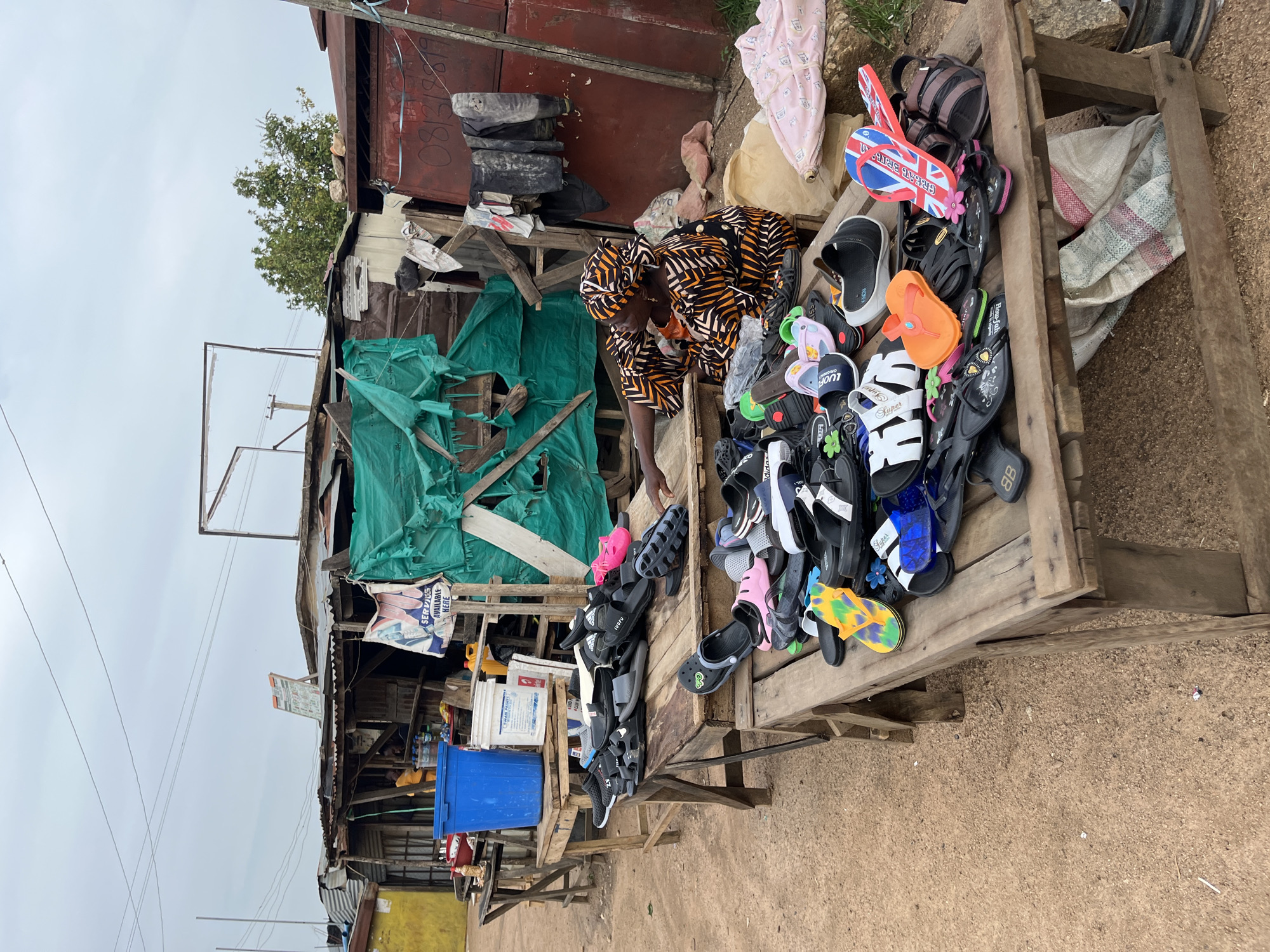 This media file captures a street vendor in Gura Top, Nigeria, selling a variety of footwear from a makeshift wooden stall. The primary subject is a woman, partially visible, wearing a patterned dress and headwrap, seemingly attending to her merchandise. The stall itself is laden with an assortment of sandals and flip-flops, displayed on a wooden table and some stacked higher. Among the footwear, notable items include a pair of flip-flops with a Union Jack design, and others with decorative elements like flowers and colorful patterns.
The background reveals a somewhat dilapidated setting, typical of a market environment. There are structures made of corrugated metal and wood, with tarps or sheeting providing cover. Some stacked tires are visible, and bags of unknown contents rest near the stall. The ground appears to be dirt or gravel. Electrical wires are also present in the upper part of the scene, suggesting an outdoor, possibly informal, commercial area.
The weather appears overcast, with a bright sky. The overall impression is that of a busy marketplace, likely in the daytime, where everyday goods are sold. The presence of the vendor and the array of products suggests economic activity. Visible text includes "SWAN PAINTS", "SERVIS AVAILABLE HERE", and brand names like "Adidas", "LUOFU", and "8B" on some of the items. There are also some numbers that appear to be a phone number: "08/312819".