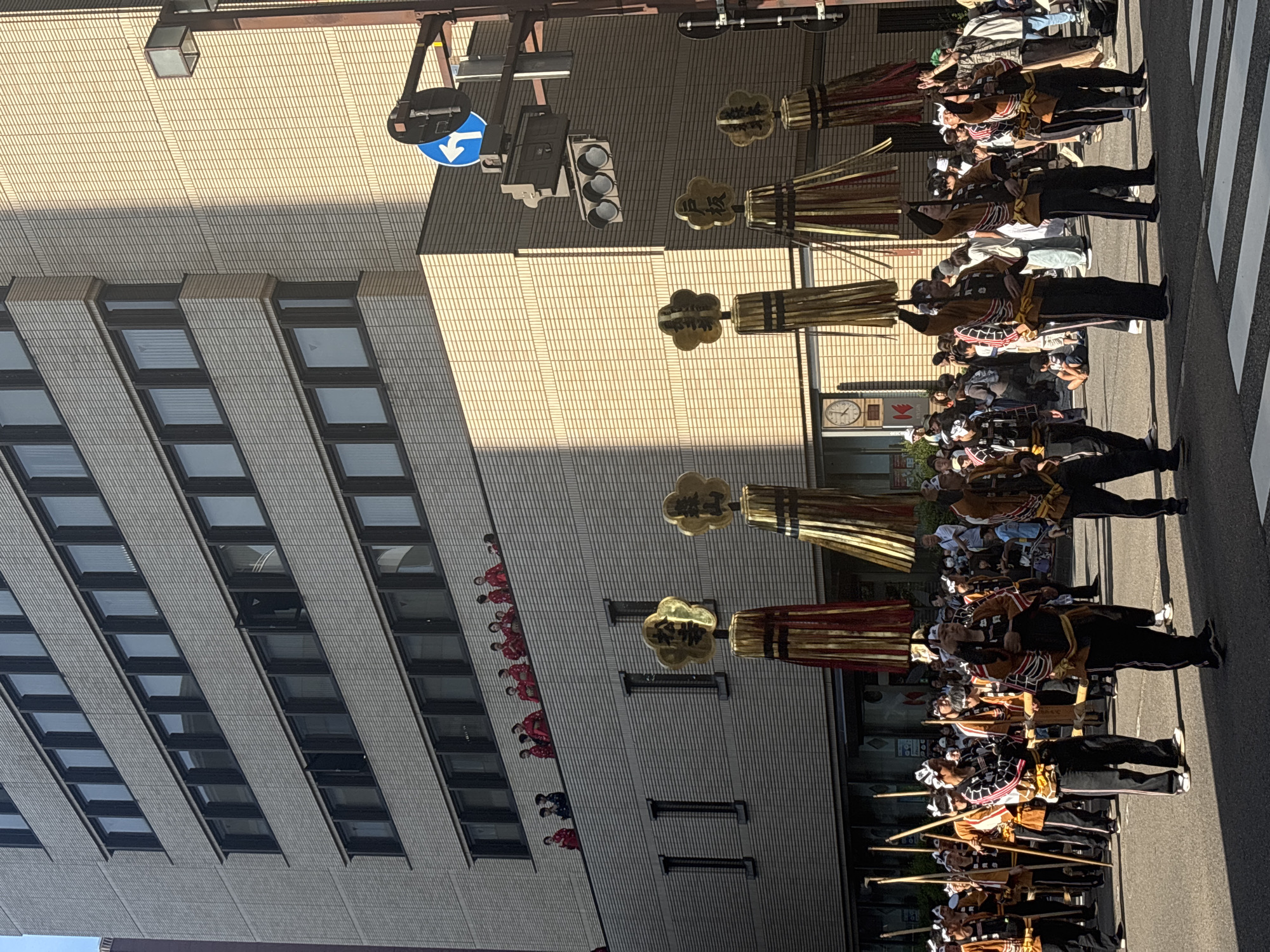 This daytime image depicts a street scene during a festival parade in Kanazawa, Japan, likely the Hyakumangoku Matsuri. The perspective is slightly elevated, looking down a city street towards a multi-story building.
In the foreground and mid-ground, a procession of approximately 20-30 individuals, predominantly men, advances on a paved street. They are dressed in traditional festival attire consisting of black trousers, short-sleeved jackets (some brown with black stripes, others patterned), and white headbands. Several men carry long wooden poles. Prominently featured are five tall, decorative elements, each topped with a gold, five-petaled motif displaying kanji characters. From left to right, these read "松ヶ枝" (Matsugae), "祭山" (Matsuyama), "祭山" (Matsuyama), "戸板" (Toita), and "松ヶ枝" (Matsugae). Beneath these gold tops are cylindrical structures composed of vertical red and gold, or gold and black, tassels or strips. The individuals holding these elements typically have them raised above their heads or shoulders. Spectators line the sides of the street behind the procession. The street surface includes a visible white crosswalk in the lower right.
In the mid-ground, on a lower-level balcony or terrace of the multi-story building, a group of approximately 10-15 women is observing the parade. They are attired in red kimonos, some with dark patterns, and have traditional hairstyles.
The background is dominated by the multi-story building, which features a light-colored brick or tiled facade and multiple rows of rectangular windows with dark frames. The building exhibits recessed sections on some floors, creating architectural variation. On the right side of the image, a dark metal pole supports a set of three dark-lensed traffic lights and a circular blue sign with a white left-turn arrow. A square-shaped street light fixture is also mounted on the pole. Strong sunlight illuminates the upper right portion of the building, casting distinct shadows across the street and lower parts of the building.