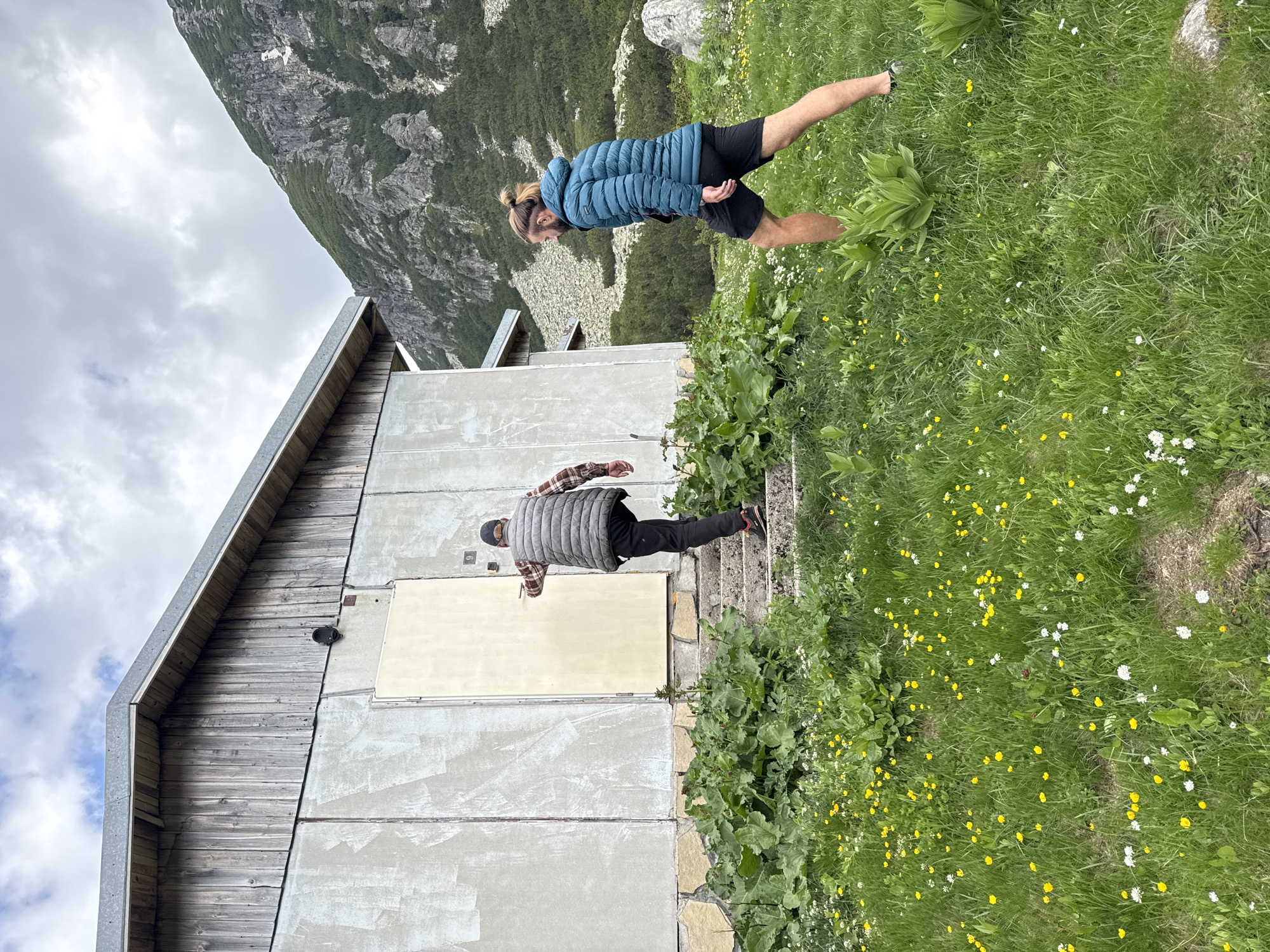 Two men are walking on a grassy slope towards a rustic building in the mountains near Yakoruda, Bulgaria. The building has a concrete facade and a wooden roof. One man, wearing a plaid shirt and a puffer vest, is climbing a set of stone steps towards a door. He is from the back and is wearing a baseball cap. The other man, wearing a turquoise puffer jacket and black shorts, is walking on the grass to the right, facing the building. The surrounding environment is mountainous with rocky terrain and vegetation, including green grass and yellow wildflowers. The sky is cloudy.