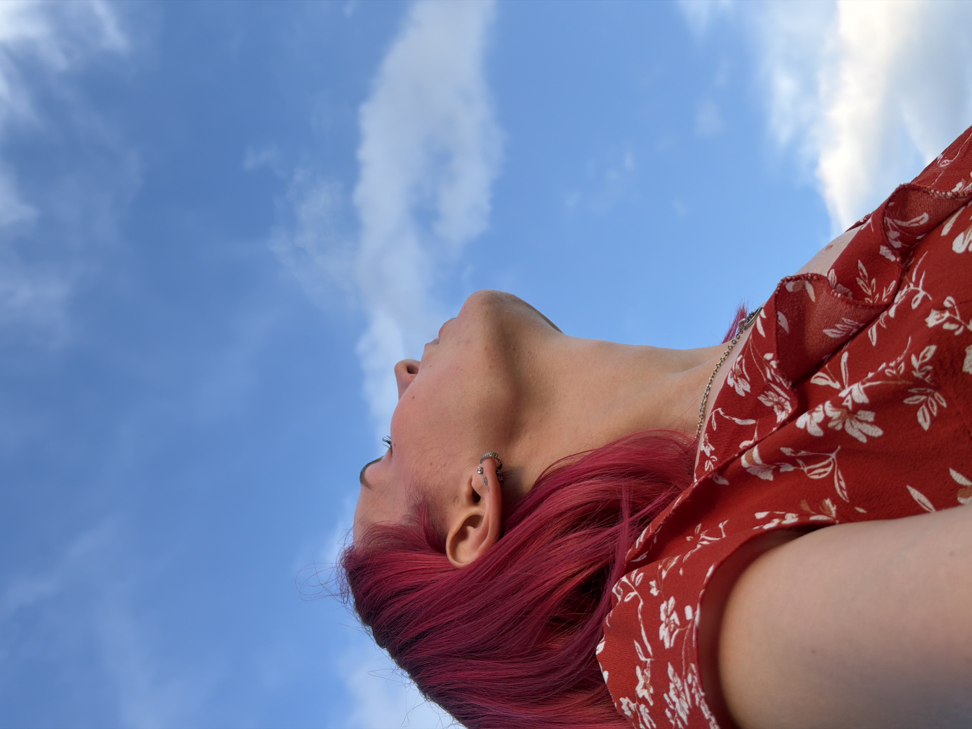 A close-up, low-angle shot captures a person with vibrant pink hair looking up into a bright blue sky with scattered white clouds. The person is wearing a red dress with a white floral pattern. They have piercings in their ear, including a hoop earring and smaller studs. The image is likely from Chaikovsky, Russia, given the bright, clear sky and the aesthetic. The lighting suggests it's daytime.