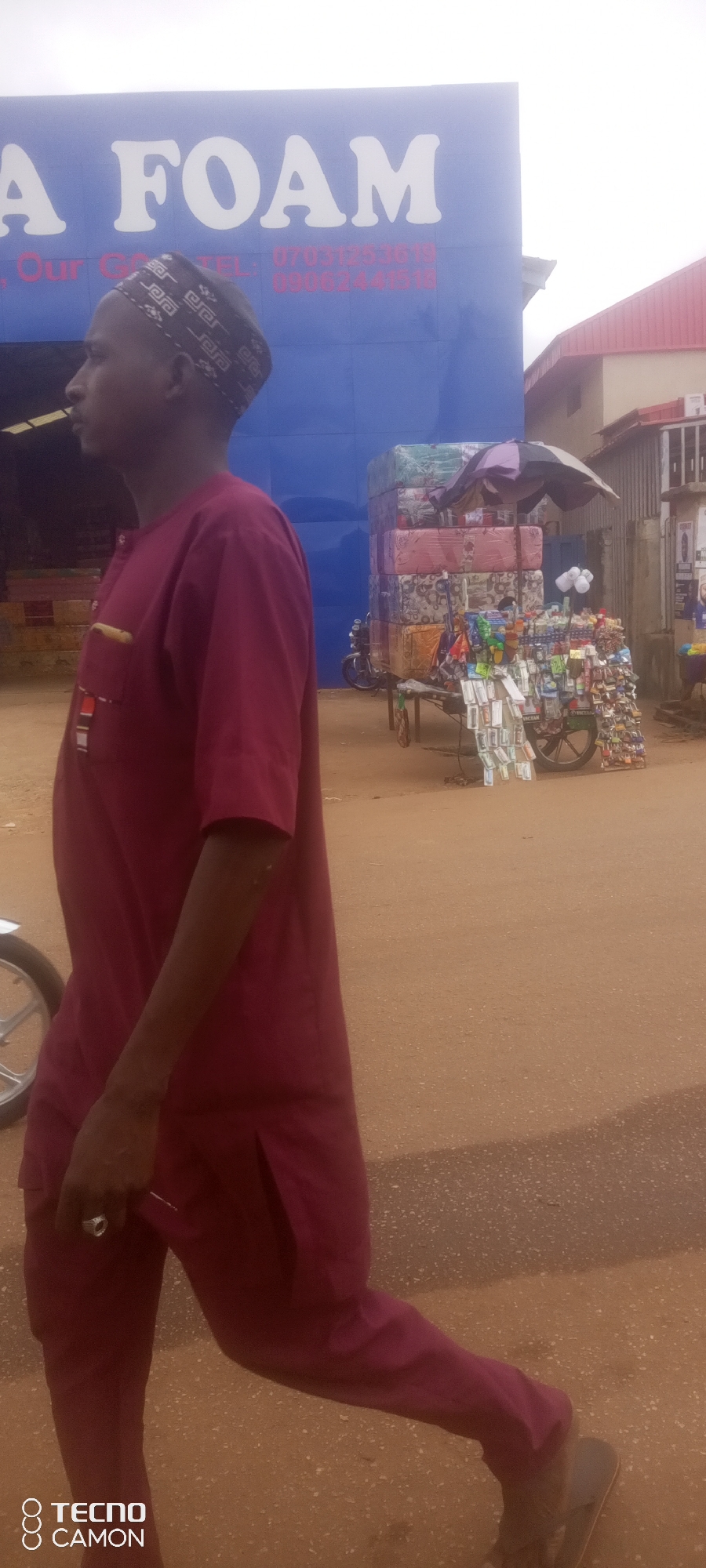 A man in a maroon traditional outfit and cap walks across a dirt road in front of a blue building that has "A FOAM" printed in large white letters. The building also displays smaller red text that includes phone numbers, likely for the business. To the right of the man and in front of the building, a multi-tiered cart is laden with stacked mattresses and various small items displayed for sale. A motorcycle is parked behind the cart, and further to the right, there are buildings with red roofs. The sky is overcast. The scene appears to be in Ankpa, Nigeria.