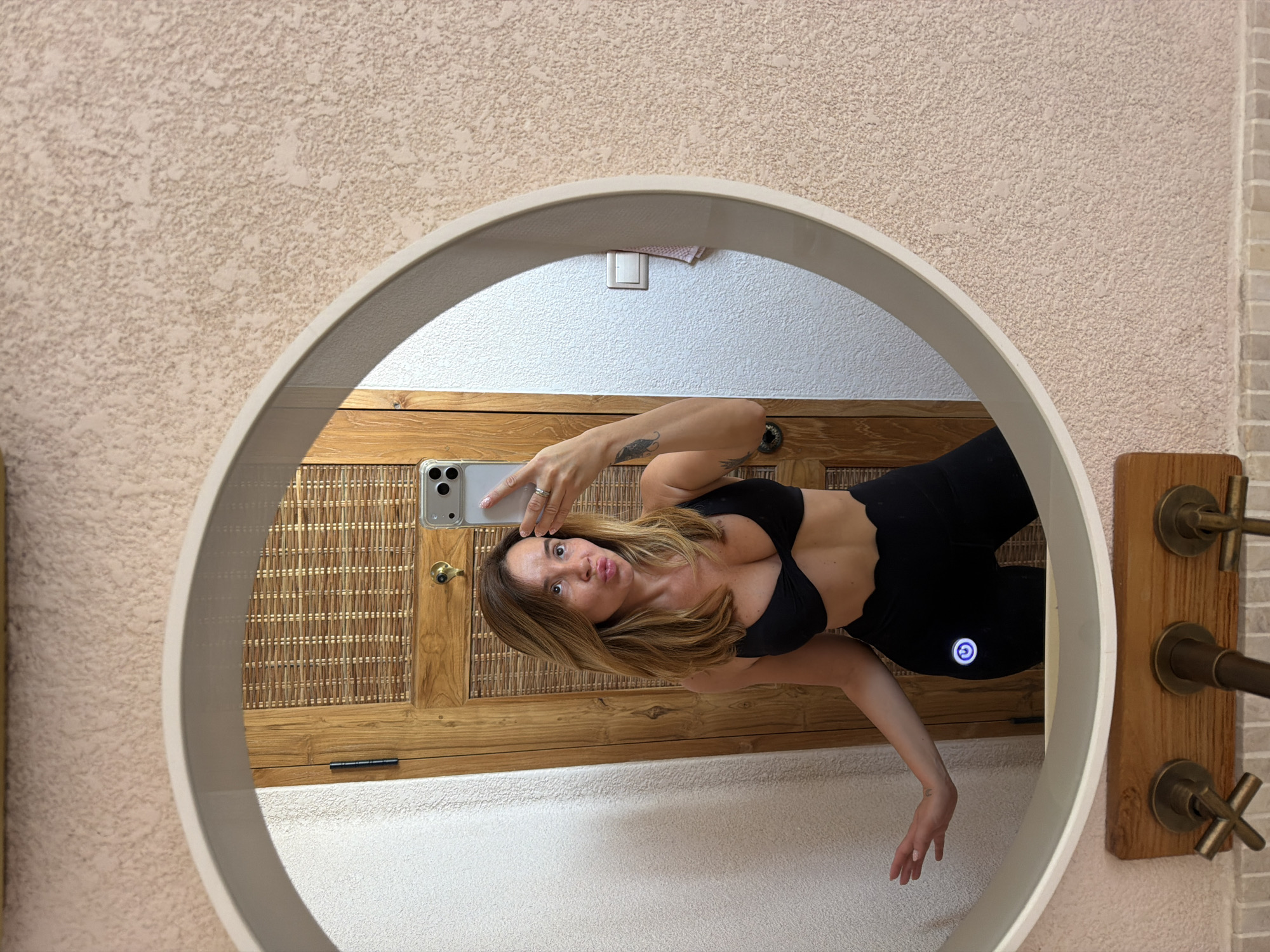 A woman in a black bikini top and black leggings is taking a selfie in a round mirror. She is standing in a room with textured walls and a wooden door behind her. The mirror reflects her and part of the room. She is looking at the phone and puckering her lips. She is wearing a ring on her right hand. There is a tattoo on her left arm. The leggings have a glowing circular logo on the left thigh. The bathroom has brass fixtures. The overall ambiance suggests a tropical or resort setting. The specific location within Mengwi, Indonesia cannot be determined from the visual information provided.
