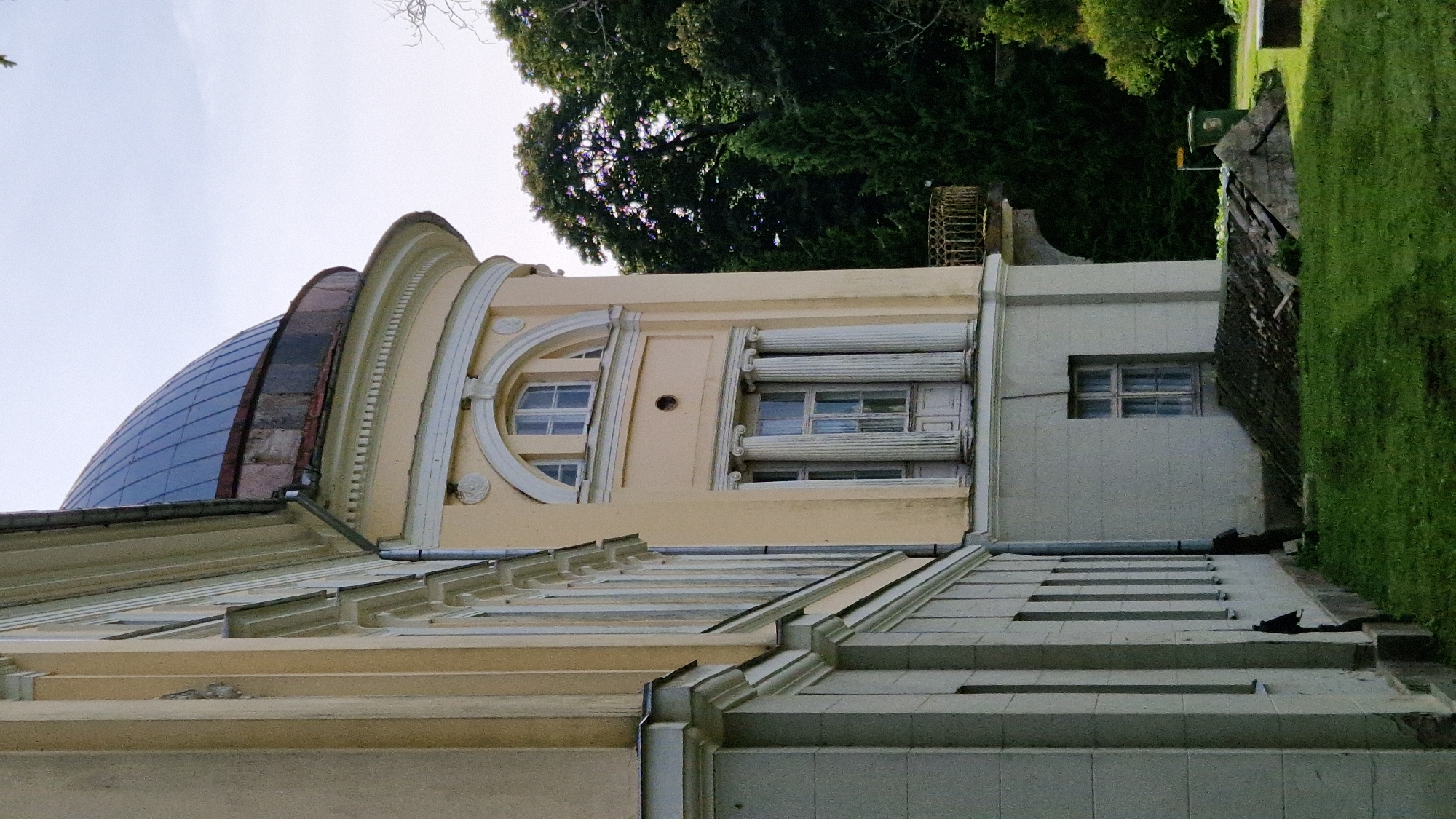 The image shows a side view of a light yellow classical building with a blue domed roof. The building has a complex facade with arched windows, columns, and decorative molding. To the right of the building, there are several mature trees, some of which are evergreen. In the foreground, there is a grassy area with some construction debris or landscaping materials. The sky is overcast. The building appears to be part of the Kazdanga Manor complex in Kazdanga, Latvia. There are no people visible in the image.