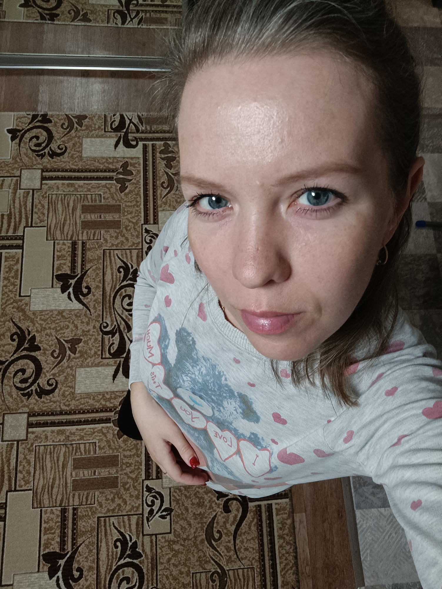 A woman with fair skin, blue eyes, and light brown hair pulled back from her face is pictured from a high angle. She is wearing a light gray long-sleeved shirt with pink heart patterns and a graphic featuring the word "LOVE." The woman is wearing small, gold hoop earrings. Her fingernails are painted red on her left hand which is visible.
The woman is standing on a floor with a patterned brown and beige covering. The pattern includes floral motifs and geometric shapes.
The photograph appears to be taken indoors. There is no visible information to determine it was taken in the city of Chaikovsky, Russia.