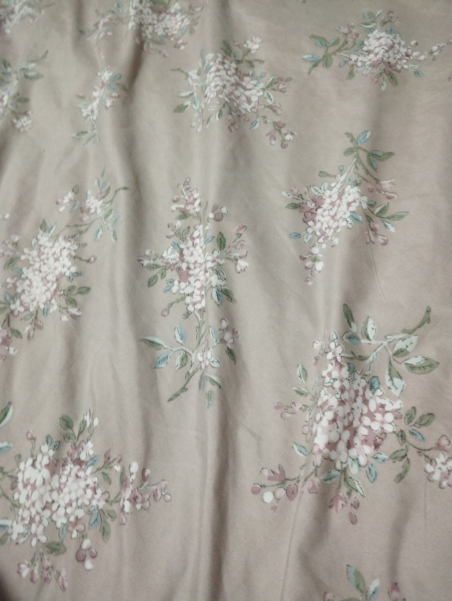 This is a photograph of a fabric with a floral pattern. The fabric is a light beige color and features clusters of small white and pink flowers with green leaves. The flowers are arranged in a scattered pattern across the fabric. The image is a close-up shot, and there are visible wrinkles and folds in the fabric, suggesting it might be a bedsheet or curtain. There are no people or specific landmarks visible. The location "Manaskent, Russia" is not depicted in this image.