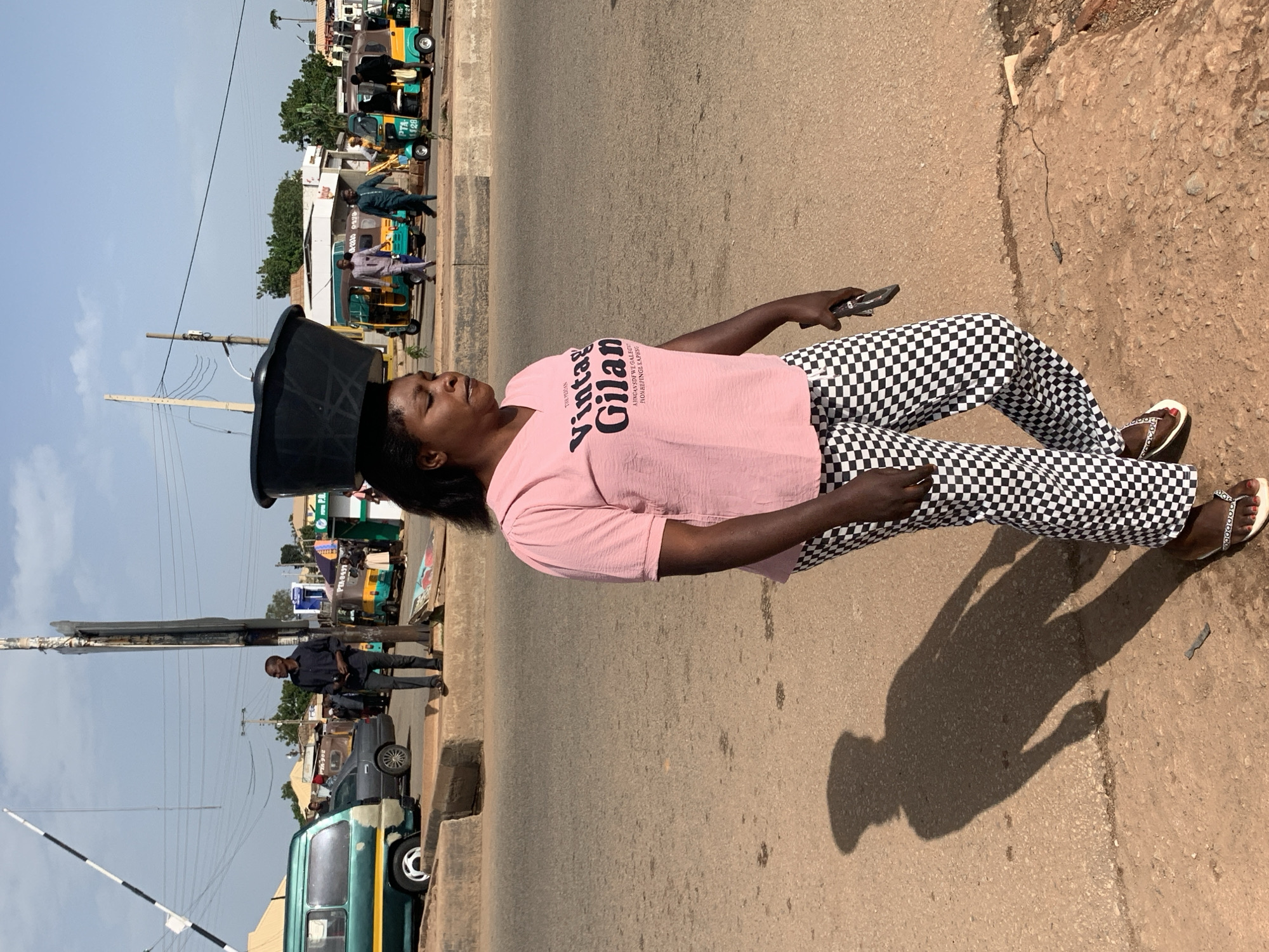 A woman with dark skin is walking on a dusty road in Gura Top, Nigeria, balancing a large black tub on her head. She is wearing a pink t-shirt with black lettering and black and white checkered pants. She is also wearing flip-flops. A shadow of the woman is cast on the road behind her. In the background, there are several vehicles, including a green bus, some auto-rickshaws, and a few people standing around. The sky is blue with some clouds. The scene appears to be a busy street in a marketplace or transportation hub.