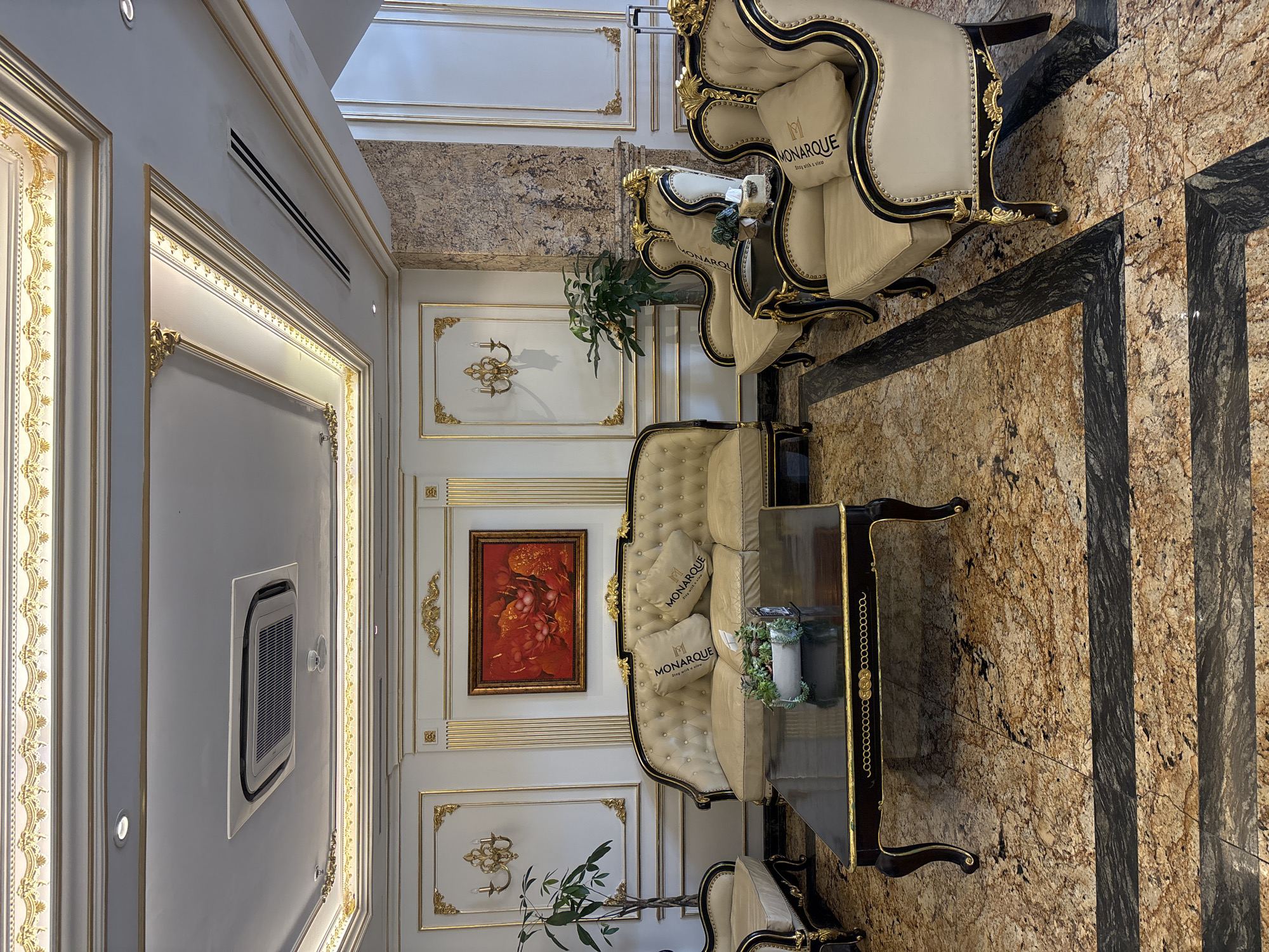 The image depicts a luxurious hotel lobby in Da Nang, Vietnam, featuring ornate decor and elegant furnishings. The room is characterized by its high ceilings with intricate gold trim and recessed lighting. A prominent air conditioning unit is visible on the ceiling. The walls are adorned with white paneling and detailed gold accents, framing a vibrant red painting of lotuses.
Seating in the lobby includes a plush, cream-colored three-seater sofa and two matching armchairs, all featuring tufted upholstery and elaborate gold trim. Decorative pillows with the "MONARQUE" logo are placed on the sofa. A dark, rectangular coffee table with gold detailing sits in front of the sofa, holding a small potted plant and a tissue box. A single armchair with similar styling is partially visible to the left.
The floor is covered in a polished marble with a golden-brown granite pattern, contrasted by dark horizontal bands. A large potted plant stands to the left of the sofa, and another plant is visible near the armchairs. The overall impression is one of opulence and refined taste, consistent with a high-end hotel environment in Da Nang.