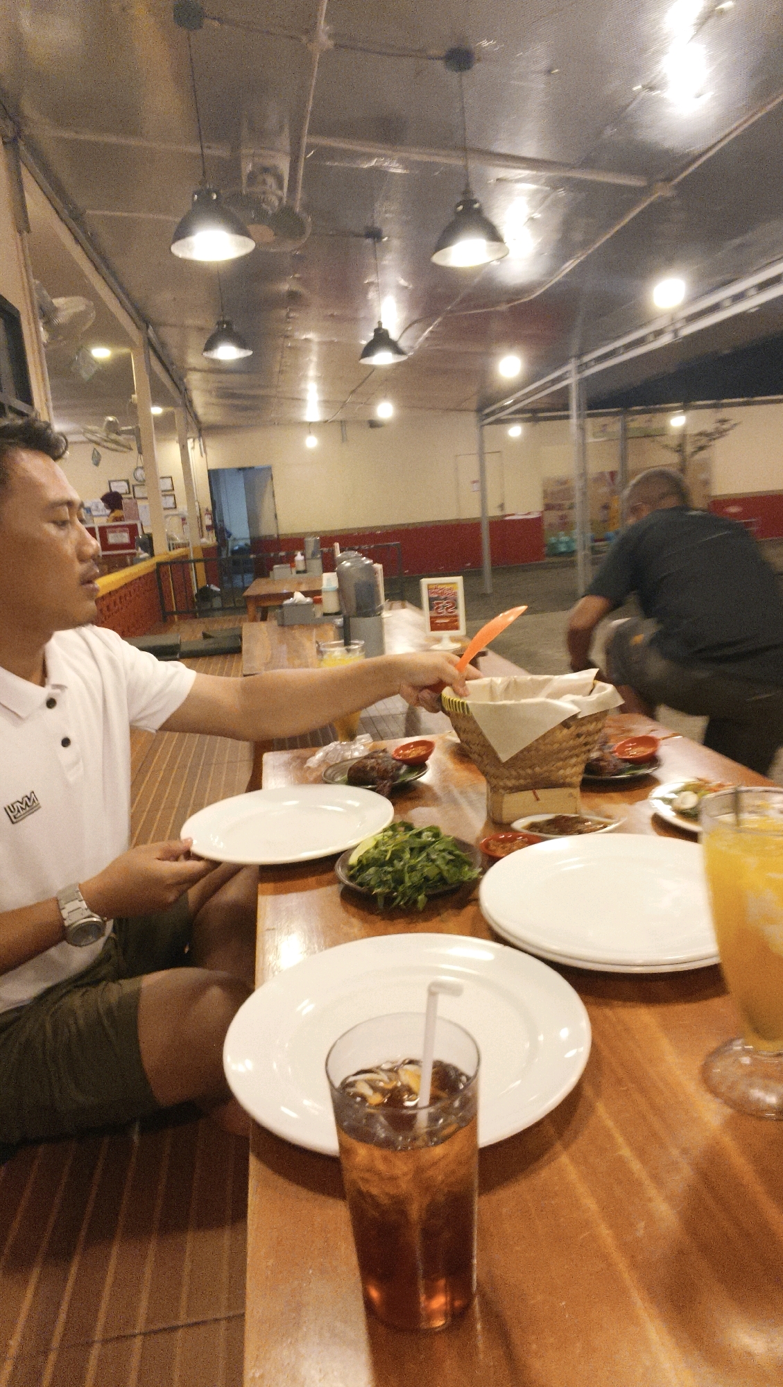 The image captures an indoor dining scene, likely a restaurant, set at night. The ambient lighting comes from overhead industrial-style pendant lamps, casting a warm glow on the metallic ceiling and wooden tables. The setting appears to be in Ketapang, Indonesia, indicated by visual cues and context.
In the foreground, a man wearing a white polo shirt with a logo, olive green shorts, and a wristwatch is seated at a wooden table. He is holding a white plate and reaching with his right hand towards an orange utensil protruding from a woven basket. Before him are several plates, a small bowl of green vegetables, and a glass filled with iced tea, complete with a straw.
To the right, the upper portion of another person is visible, seated and holding a glass containing an orange beverage, possibly juice. In the background, another individual, seen from the back, is bent over near a table, appearing to be engaged in some activity. The restaurant's interior features additional wooden tables and chairs, along with exposed ceiling structures and lighting. The overall atmosphere suggests a casual dining experience.