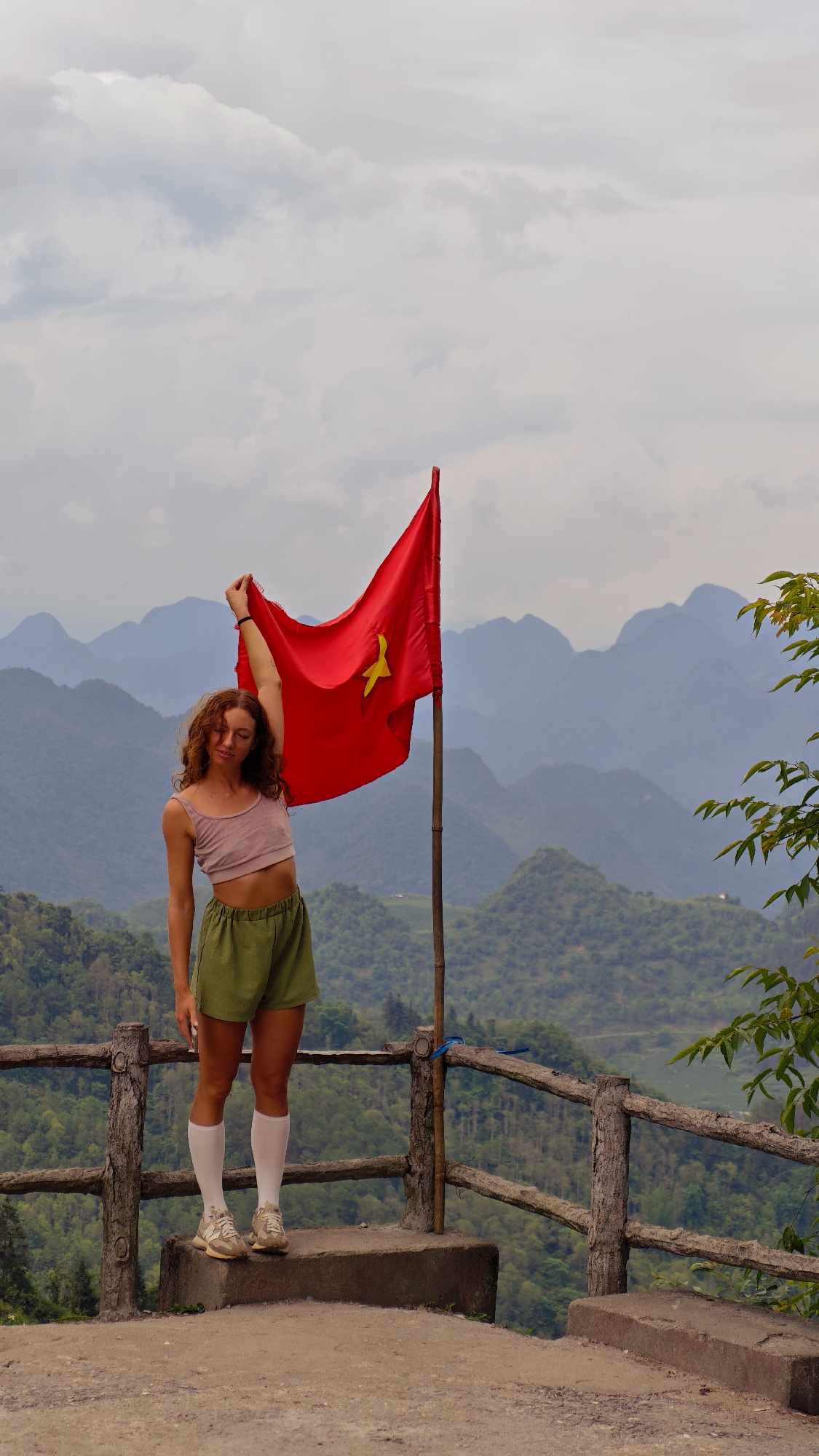A young woman stands on a concrete platform overlooking a mountainous landscape in Tam Son, Vietnam. She holds the Vietnamese flag aloft with her right hand, its red color and yellow star prominently displayed. The woman wears a crop top, green shorts, and white socks with athletic shoes. A rustic wooden fence lines the edge of the platform, offering a view of the verdant, tiered mountains in the background, obscured by a cloudy sky.