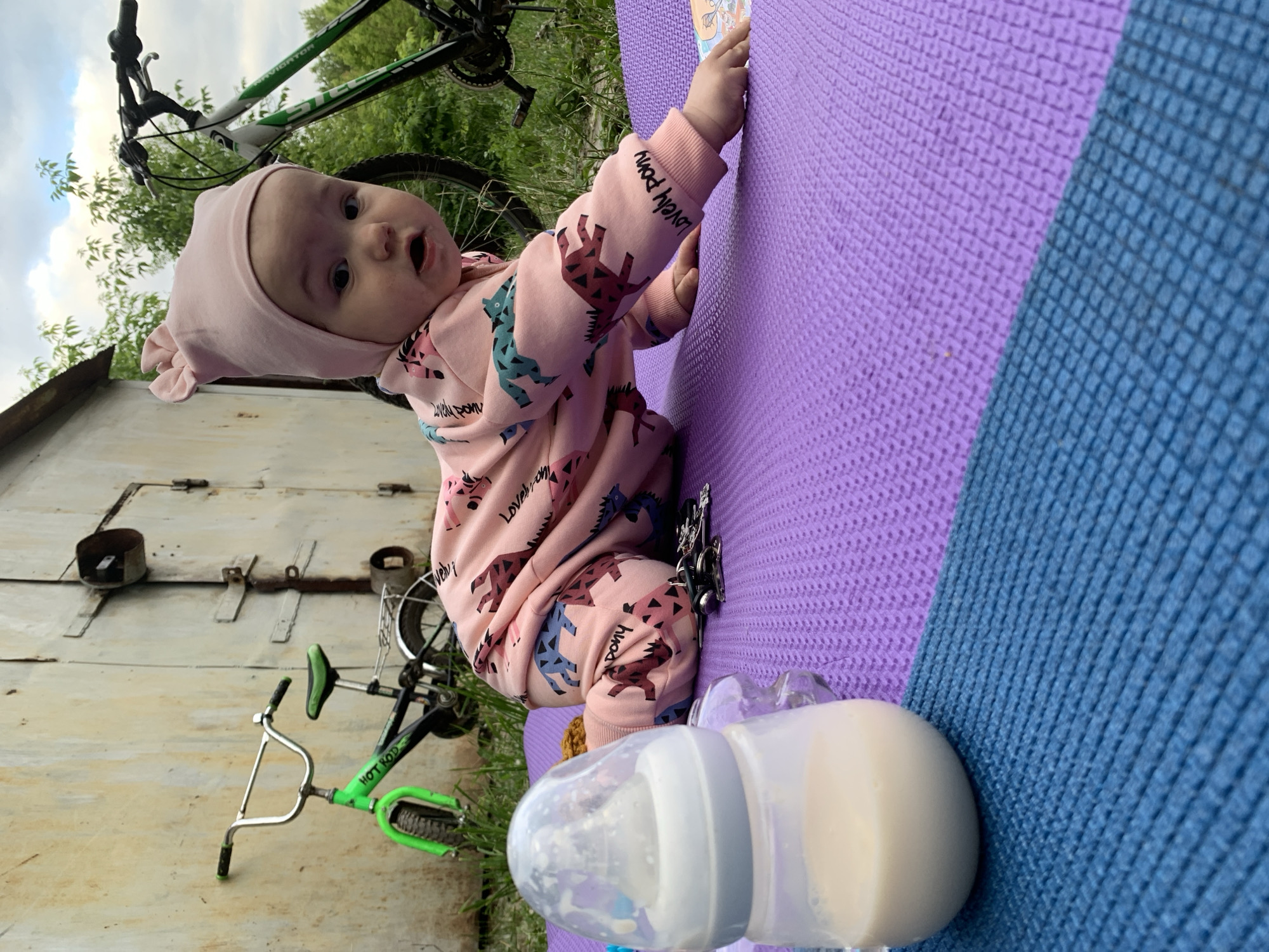 A baby in a pink onesie with a bunny-ear hat is crawling on a purple mat in front of a corrugated metal garage door in Chaikovsky, Russia. The baby is looking at the camera with an open mouth. Next to the baby is a bottle of milk. In the background are two bicycles. One is a child's bike and the other is a mountain bike. The grass is green and there are trees in the background. The sky is cloudy.