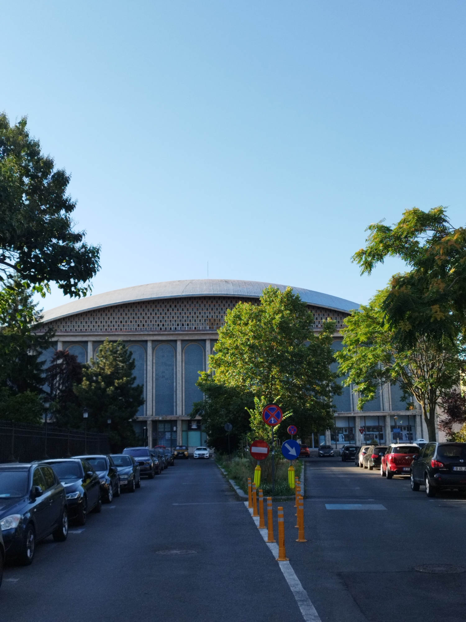 The image depicts the exterior of the Sala Palatului in Bucharest, Romania, on a clear, sunny day. The large, oval-shaped building is partially obscured by lush green trees on either side and in front of it. The building's facade features a repeating pattern of square tiles above a section with tall, arched windows. A light-colored, domed roof caps the structure.
In the foreground, a street runs away from the camera, lined with parked cars on both sides. Several traffic signs are visible on the left side of the street, including a "no stopping" sign and a directional arrow sign. Orange traffic cones are also placed along the edge of the street. The cars are of various makes and models, parked in a single file on the right side of the street. The sky above is a clear, pale blue.
