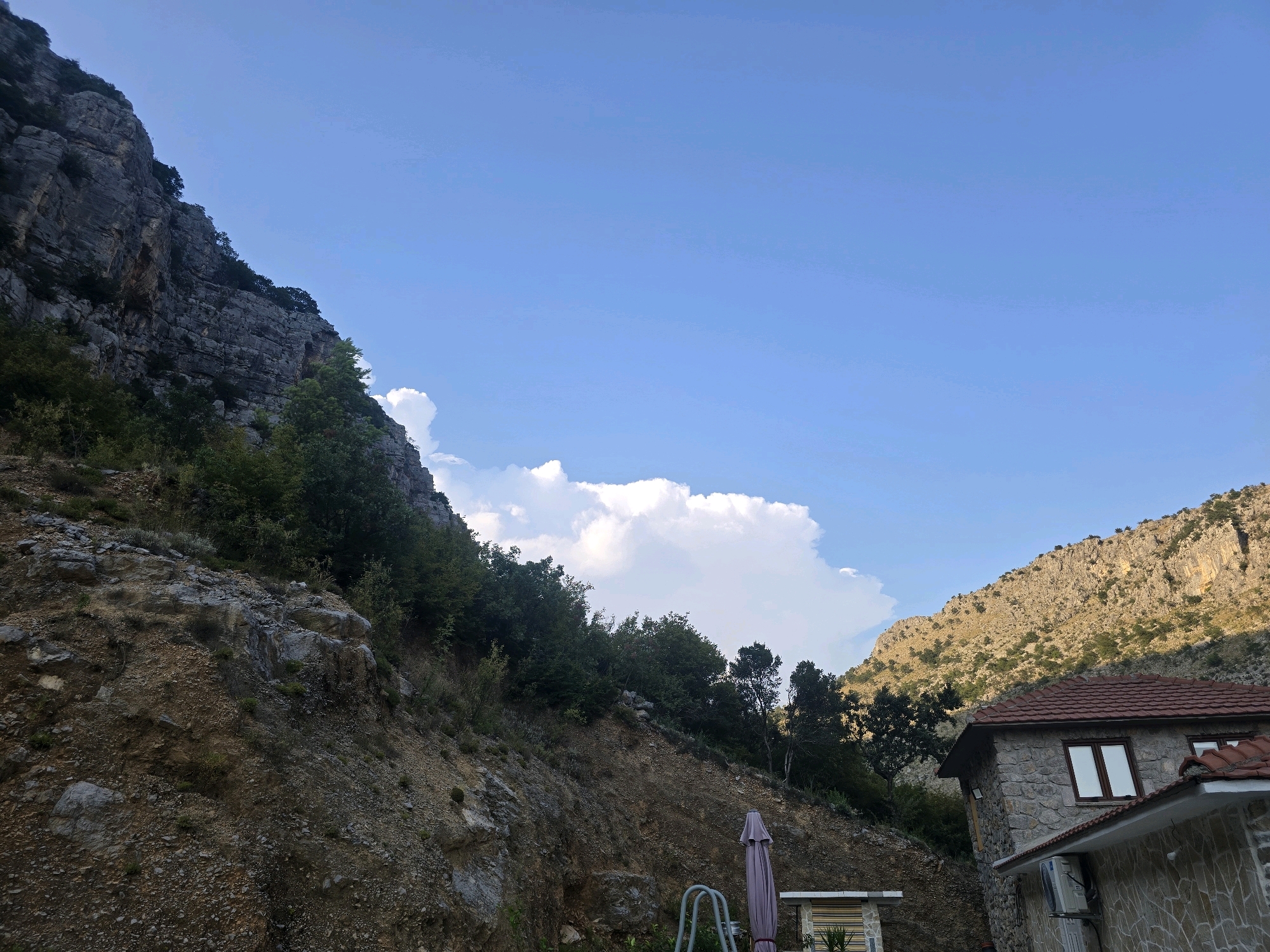 This image depicts a mountainous landscape with a clear blue sky. On the left side of the frame, a steep, rocky cliff face is visible, covered in sparse vegetation. To the right, a stone building with a red tiled roof is situated, featuring windows and an air conditioning unit mounted on the exterior wall. A closed umbrella and part of a metal railing are also present in the foreground. The terrain appears to be uneven and inclined, with soil and rocks dominating the ground. The scene suggests a rural or semi-rural setting within or near Podgorica, Montenegro, characterized by its proximity to natural mountainous terrain and a residential structure.