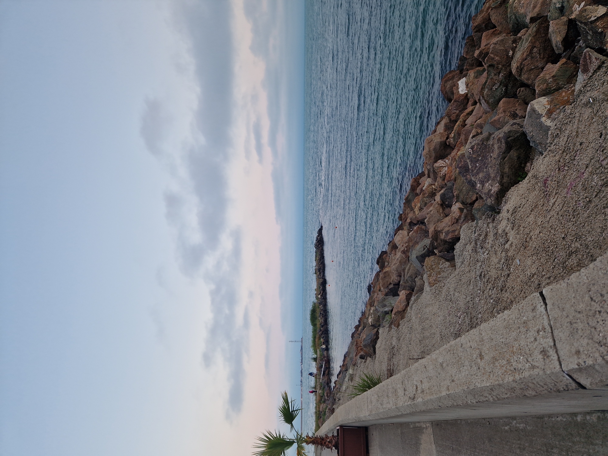 The image captures a coastal scene during twilight or an overcast day, presenting a view of a tranquil sea and a rocky shoreline. The sky above is a pale blue-grey, predominantly covered by scattered, light-colored clouds, with a clearer, lighter expanse at the top. The horizon line is slightly hazy, blending the sky into the distant water.
The sea, occupying the right half and extending towards the horizon, is a deep blue-green, exhibiting a smooth surface with minimal ripples, suggesting calm conditions. Several small, orange, spherical buoys are visible floating in the mid-distance water.
In the foreground, a concrete path runs along the left side of the frame, separated from the rocky shore by a low, weathered concrete barrier or wall. This barrier shows a distinct crack near its center in the immediate foreground. To the left, a small palm tree in a dark planter box sits on the concrete path.
The immediate shoreline consists of numerous large, irregular, brownish-grey and reddish-brown rocks, forming a natural or constructed breakwater. Further along the shore, a narrow, vegetated landmass extends into the sea, appearing as a small peninsula or jetty. Two human figures are visible on this landmass: one wearing a pink top, the other a blue top, both standing near a small wooden platform or structure.
In the far background, across the water, a cluster of indistinct structures, possibly indicative of a port or industrial area, can be seen. A prominent, tall, slender structure, resembling a lighthouse or a communication tower, stands out against the faint horizon in the distant center-left. The overall lighting is soft and diffused, creating a serene atmosphere.