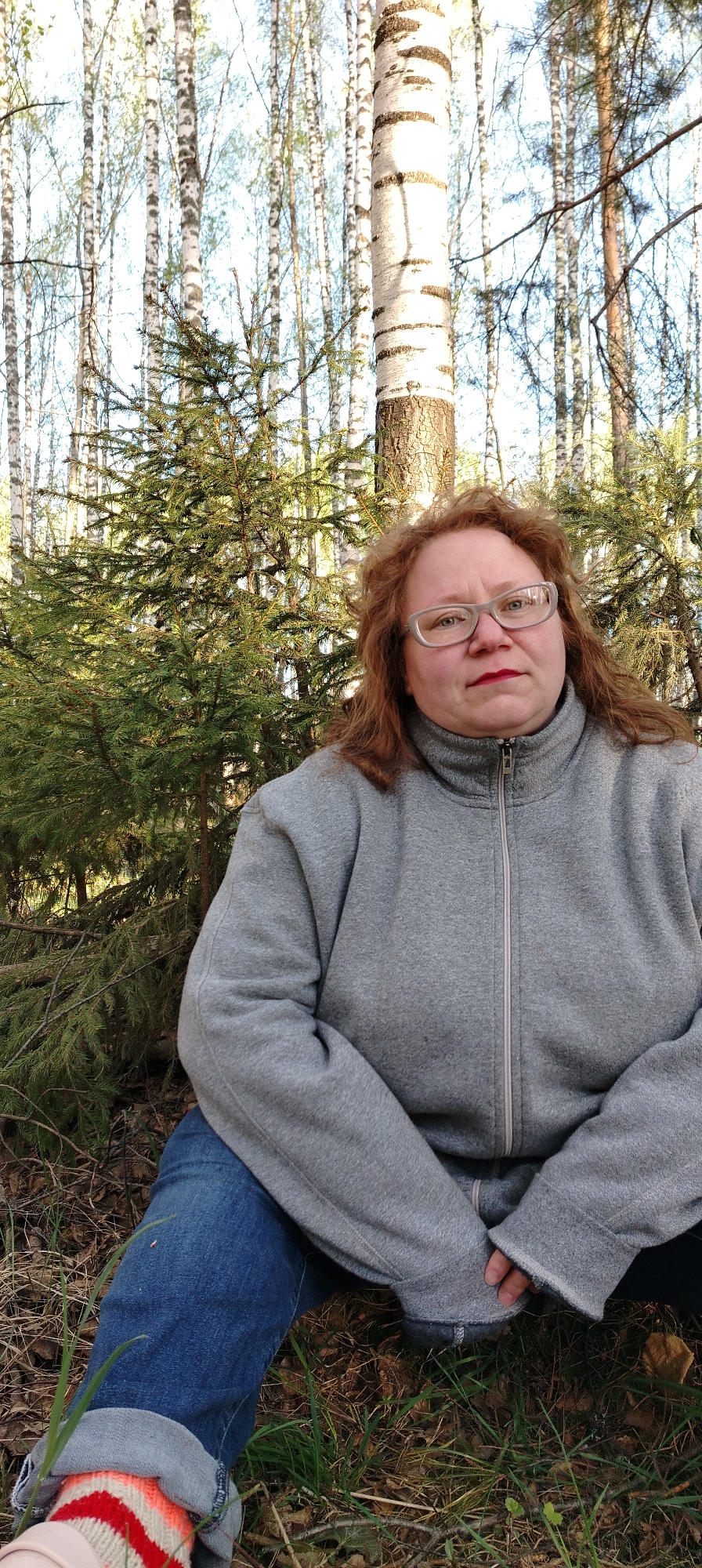 The image shows a woman with reddish-brown hair and glasses, wearing a gray zippered fleece jacket and blue jeans, sitting on the ground in a forest or wooded area. The location is Zipunovo, Russia. Behind her are several birch trees and a small evergreen tree. The ground is covered with fallen leaves and grass. A partially visible foot with a striped sock is in the lower left corner of the image.