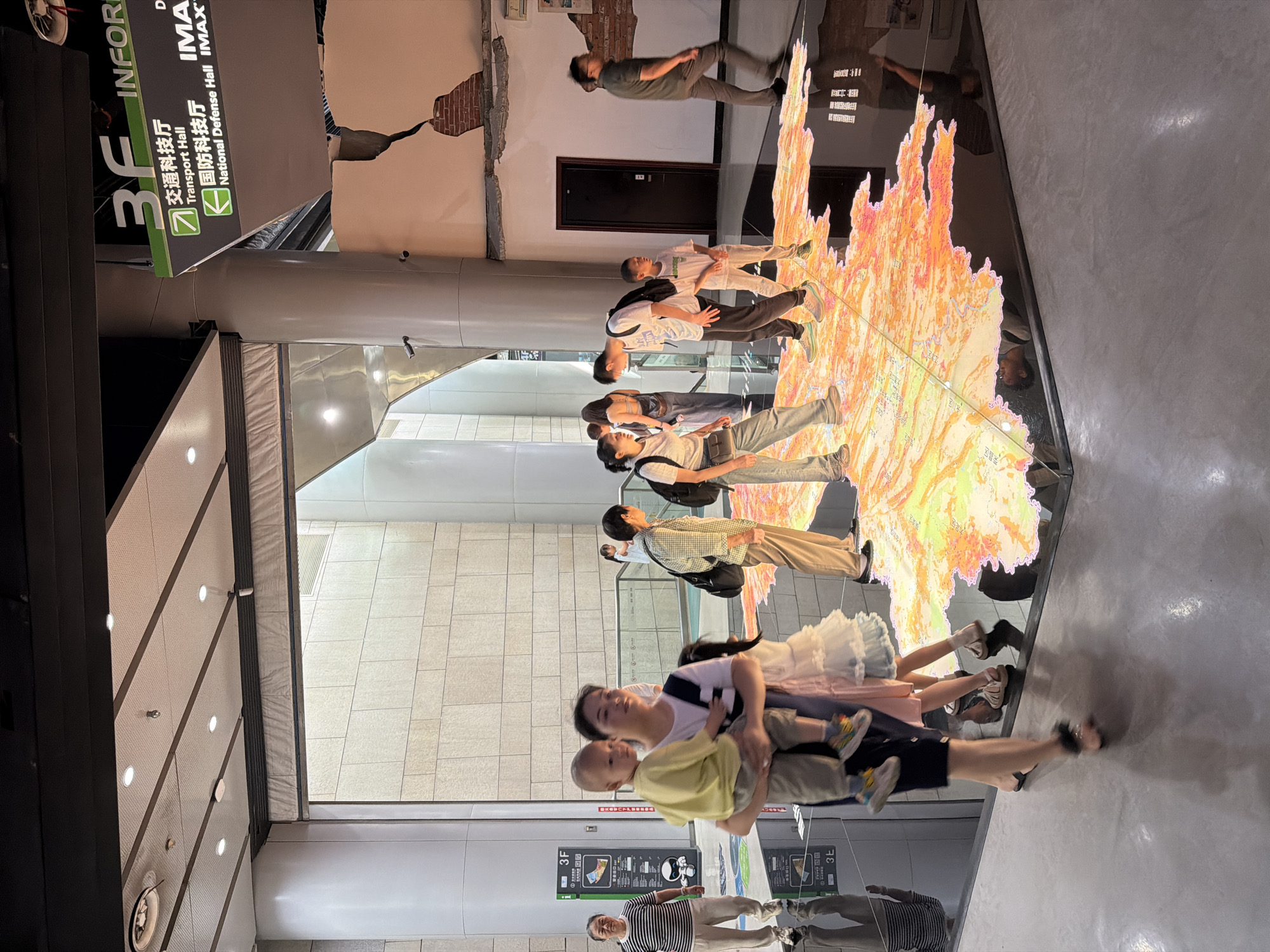 An indoor scene with a group of people interacting with a large, illuminated floor map displaying topographical and cultural features, likely a museum or exhibition. Several individuals, including adults and children, are standing or walking on and around the map. The map is brightly lit with orange and yellow hues, depicting what appears to be a geographical area with detailed markings. The environment includes large pillars, a textured wall with visible damage, and directional signage indicating "3F", "Transport Hall", and "National Defense Hall IMAX". The flooring is dark and reflective, mirroring the illuminated map. The people are casually dressed, suggesting a public and accessible space. The overall atmosphere appears to be one of exploration and engagement with the exhibit. While the specific location within 江北城街道, China is not explicitly stated, the presence of Chinese signage and the overall architectural style suggest an urban setting in China.