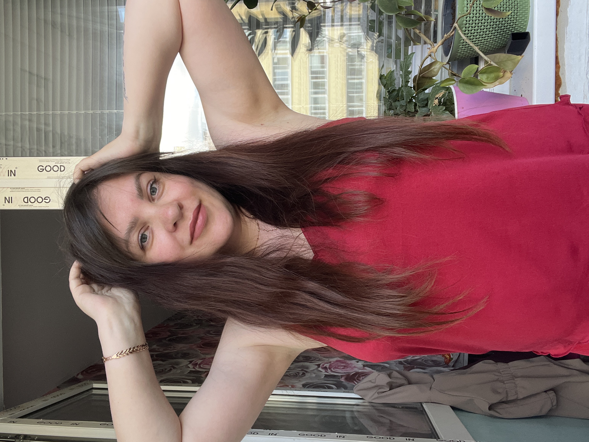 The media file shows a person posing for a selfie, holding one hand behind their head and the other resting on their arm. They are wearing a red top and a bracelet. The background appears to be a balcony or window area, with vertical blinds and a potted plant visible. Outside the window, there are buildings with yellow facades and white windows, suggesting an urban setting. The lighting indicates it might be daytime. The person has long brown hair and is looking at the camera with a slight smile.
Visible text includes "GOOD IN" repeated on what appears to be a box or packaging in the lower part of the image. The location context is Noviy, Russia.