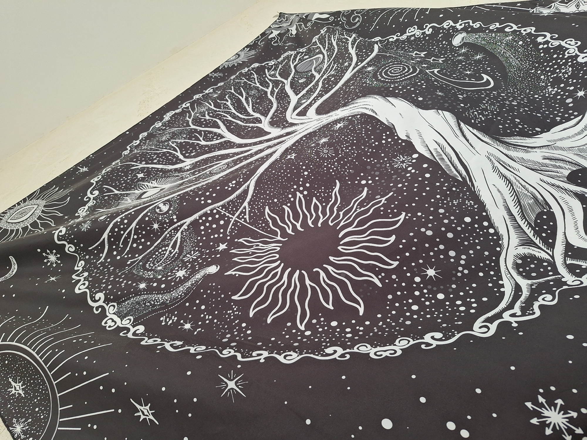 The image shows a dark fabric with a white print featuring celestial and tree-like imagery. Dominating the design is a large tree with bare branches reaching across the top and left side. To the lower left is a sun graphic. The fabric is adorned with white dots, likely stars, suggesting a night sky. The edge of the fabric appears to meet a white wall. There is no visible indication that the image was taken in Panagyurishte, Bulgaria.