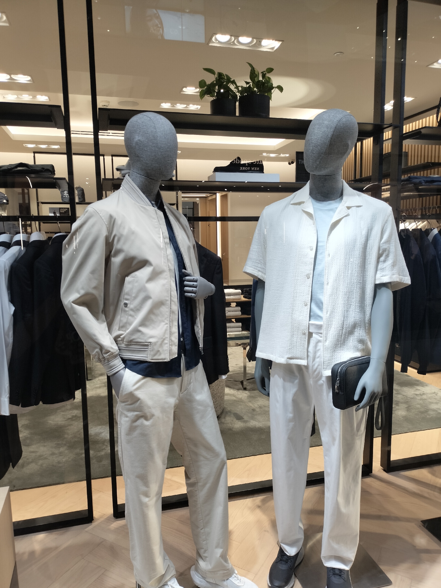 This is an indoor photograph of a retail clothing display featuring two male mannequins. Both mannequins are positioned centrally within the frame and are dressed in light-colored casual wear. The mannequin on the left wears a cream-colored bomber jacket over a navy blue collared shirt and cream-colored pants. Its left hand is tucked into its pants pocket. The mannequin on the right is dressed in a white short-sleeved, textured collared shirt over a light blue t-shirt and white pants. It holds a black rectangular pouch in its right hand. The mannequins stand on a light wooden floor.
Behind the mannequins, the background shows a retail store environment with shelves displaying clothing and accessories. Various racks of clothing, some black and some blue, are visible on the right side of the image. In the upper section of the image, there are plants in black pots and reflections from overhead lighting and what appears to be signage. The overall lighting is bright and even, typical of a commercial retail setting. The presence of mannequins and clothing racks suggests this is a fashion store. There is no visual information in the image to confirm it is located in Surabaya, Indonesia.